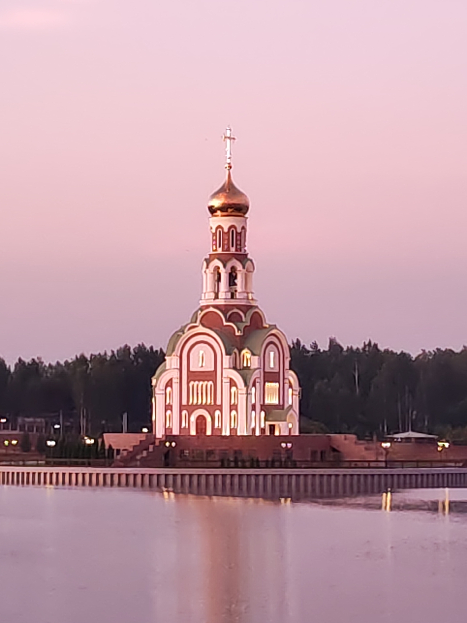 The image shows a large, ornate Russian Orthodox church with a golden dome, situated on the waterfront of the city of Sverdlovsk, Russia. The church is illuminated, casting a warm glow on its white and red facade, and its reflection is visible in the still water. A row of decorative lamps lines the walkway in front of the church, and a dense forest can be seen in the background. The sky is a soft pink, suggesting it is either sunrise or sunset. There are no people visible in the image.