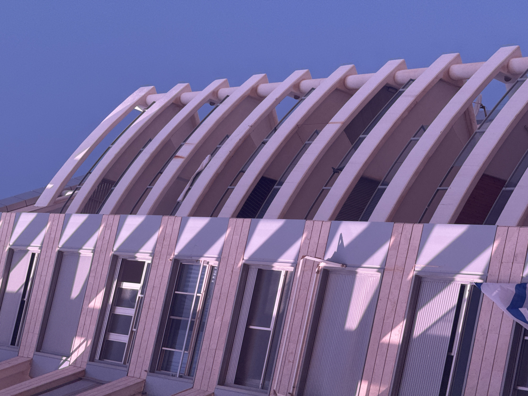 This image captures an architectural detail of a modern building in Bat Yam, Israel, under a clear, bright blue sky. The upper section of the building features a series of prominent, parallel, light-colored arched beams or ribs that form a distinctive roofline or a sun-shading structure. These beams create strong diagonal lines across the top half of the image, with gaps between them revealing darker interiors or windows.
Below this unique top section, the building's facade is visible, constructed from a light-colored material, possibly stone or concrete. It features multiple tall, rectangular windows. Long, sharp diagonal shadows, cast by the arched beams above, stretch across these windows and the facade, indicating a specific time of day, likely late afternoon or early morning, when the sun is at a lower angle. Some of the windows have venetian blinds or shutters.
In the lower right corner, a portion of the Israeli flag is visible, displaying its characteristic blue Star of David and blue stripes on a white background. The overall lighting gives the building a slightly warm, almost pinkish or purplish cast, contrasting with the vibrant blue sky. There is no visible activity or text in the image.