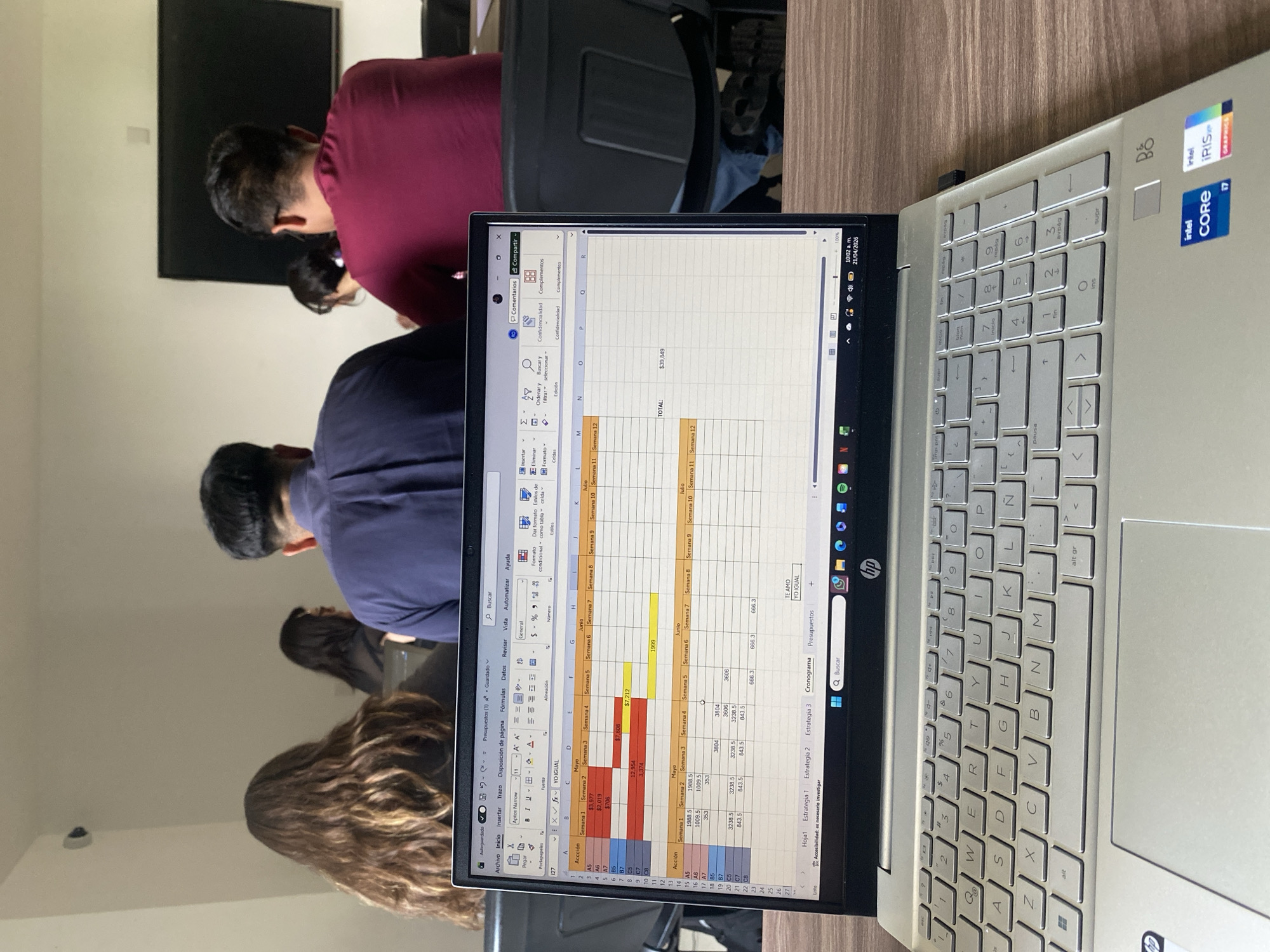 An indoor scene captured from an overhead, tilted perspective shows a silver HP laptop open in the foreground, displaying a spreadsheet on its screen. In the blurred background, several individuals are seated, facing forward, with their backs to the camera. One person wears a burgundy shirt, another a dark blue shirt, and a third has long, wavy brown hair. A black rectangular object, possibly a large monitor or television, is mounted on the white wall behind them.
The laptop screen shows Microsoft Excel with a detailed spreadsheet titled "Presupuestos (1)" (Budgets). The sheet contains columns for "Acción" (Action) and monthly breakdowns (Mayo, Junio, Julio) further subdivided into "Semana 1" through "Semana 12" (Week 1 through Week 12). Various numerical values, including monetary figures like "$3,977", "$7,608", and a "TOTAL: $39,849", are visible. Tabs at the bottom of the spreadsheet include "Hoja1", "Estrategia 1", "Estrategia 2", "Estrategia 3", "Cronograma", and "Presupuestos". A notable, informal text "TE AMO YO IGUAL" (I LOVE YOU ME TOO) appears in one cell. The top bar of the application indicates "Autoguardado" (Autosaved) and features options like "Comentarios" (Comments) and "Compartir" (Share). The laptop's system clock shows "10:02 a.m." and the date "21/04/2026". The laptop itself has an Intel Core i7 processor and Intel Iris Xe Graphics, as indicated by visible stickers. The keyboard features a Spanish layout.