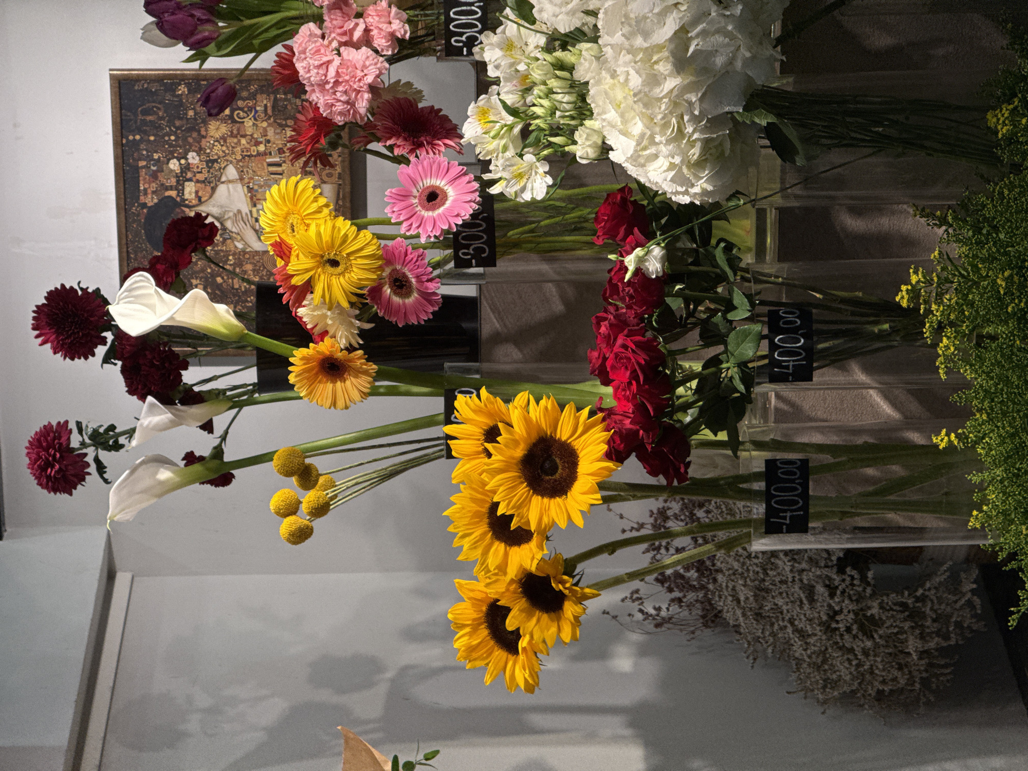 The image shows a flower shop in Belgrade, Serbia. Various cut flowers are displayed in vases, including sunflowers, gerberas, calla lilies, carnations, hydrangeas, and roses. Price tags are visible, displaying prices in Serbian currency (RSD). A framed artwork hangs in the background, potentially a reproduction of "The Kiss" by Gustav Klimt. The scene is well-lit, suggesting an indoor setting.