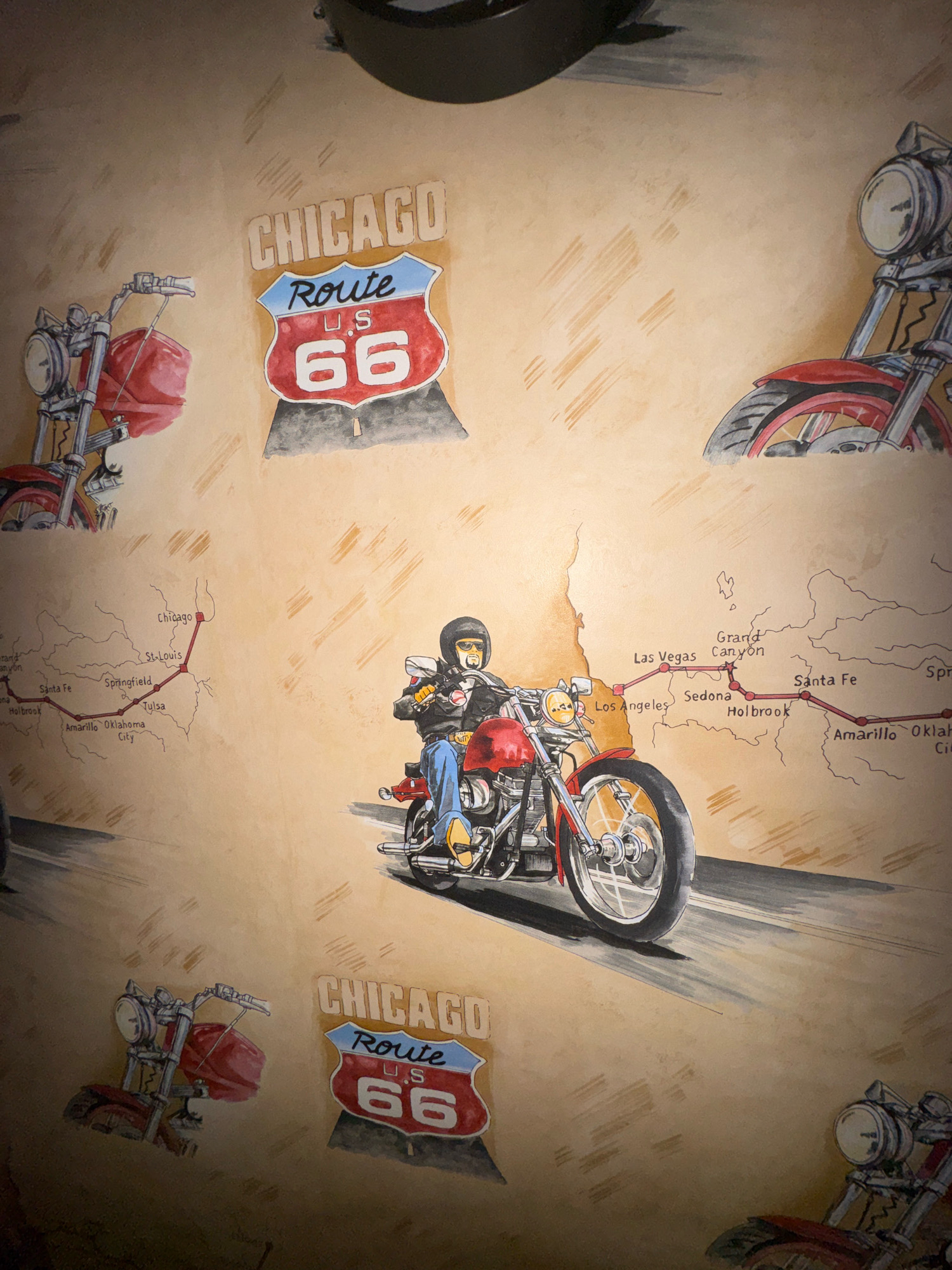 The image depicts a wall mural featuring motorcycles and a map of Route 66. The mural is rendered in a watercolor or similar artistic style, with a warm, sepia-toned background that resembles aged paper.
Several elements are visible:
* **Motorcycles:** Multiple motorcycles, predominantly red, are depicted. Some are shown in partial views, focusing on the front wheel, handlebars, and headlight. One motorcycle in the center is more fully illustrated, with a rider wearing a helmet, sunglasses, a black leather jacket, blue jeans, and yellow shoes. The rider is seated on the motorcycle, which is positioned on a road.
* **Route 66 Signage:** Two distinct Route 66 signs are present. These are the iconic shield-shaped signs, featuring "CHICAGO" at the top, "Route" in blue script, "U.S." in red, and "66" in large white numbers within a red section.
* **Route 66 Map:** A stylized map of Route 66 is integrated into the mural. It shows a red line tracing the route with various cities labeled along the way, including Chicago, St. Louis, Springfield, Tulsa, Oklahoma City, Amarillo, Holbrook, Sedona, Grand Canyon, Las Vegas, and Los Angeles. The map has a vintage, hand-drawn appearance.
* **Background and Texture:** The background is textured, mimicking parchment or an old wall, with faint, brushstroke-like lines and smudges that add to the aged effect. A dark, circular or oval shape is visible at the very top of the image, possibly a stylized representation of a motorcycle headlight or a decorative element.
The image is a close-up or medium shot, focusing on the artwork. There is no indication of a specific location in the city of Hrodna, Belarus; the content of the mural is thematic, related to American culture and road travel. The scene is static, depicting illustrations rather than a live event.