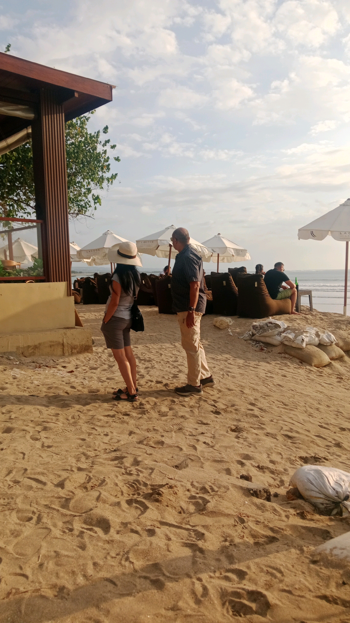A medium-wide shot shows a beach in Kuta, Indonesia, with a man and woman standing on the sand in the foreground, facing away from the camera. The man, wearing a plaid shirt and khaki pants, stands to the right of the woman. The woman, in a gray t-shirt and shorts, wears a wide-brimmed hat. Behind them, several white umbrellas with logos provide shade over beanbag chairs. To the far right, a man sits on a beanbag chair near a table with a bottle and a glass. The ocean is visible in the background, with gentle waves. Bags of sand are piled on the beach to the right. The sand is textured with footprints. The scene is illuminated by natural light, with clouds in the sky.