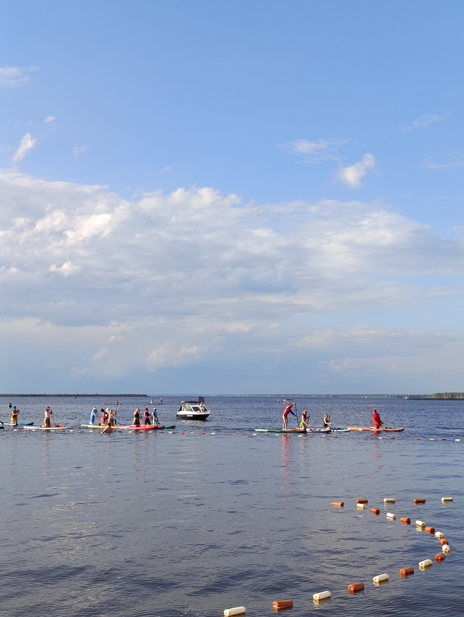 The image depicts a group of people engaged in stand-up paddleboarding on a calm body of water, likely a river or lake, under a partly cloudy sky. The scene appears to be set during the daytime, with ample natural light. In the background, a distant shore with trees and a lighthouse or similar structure is visible, suggesting a developed shoreline. A small boat, possibly a motorboat, is also present on the water to the right of the group. The water is a deep blue, reflecting the sky and clouds. The individuals on paddleboards are spread out, with some paddling and others standing still, appearing to be participating in a group activity or event. Their attire is casual, suitable for water sports. Some of the paddleboarders are wearing bright colors, adding visual interest to the scene. Scattered across the water are orange and white buoys, potentially marking a course or a designated area for the activity. The overall mood is relaxed and recreational, with a sense of community and outdoor enjoyment. The location context is Chaikovsky, Russia.