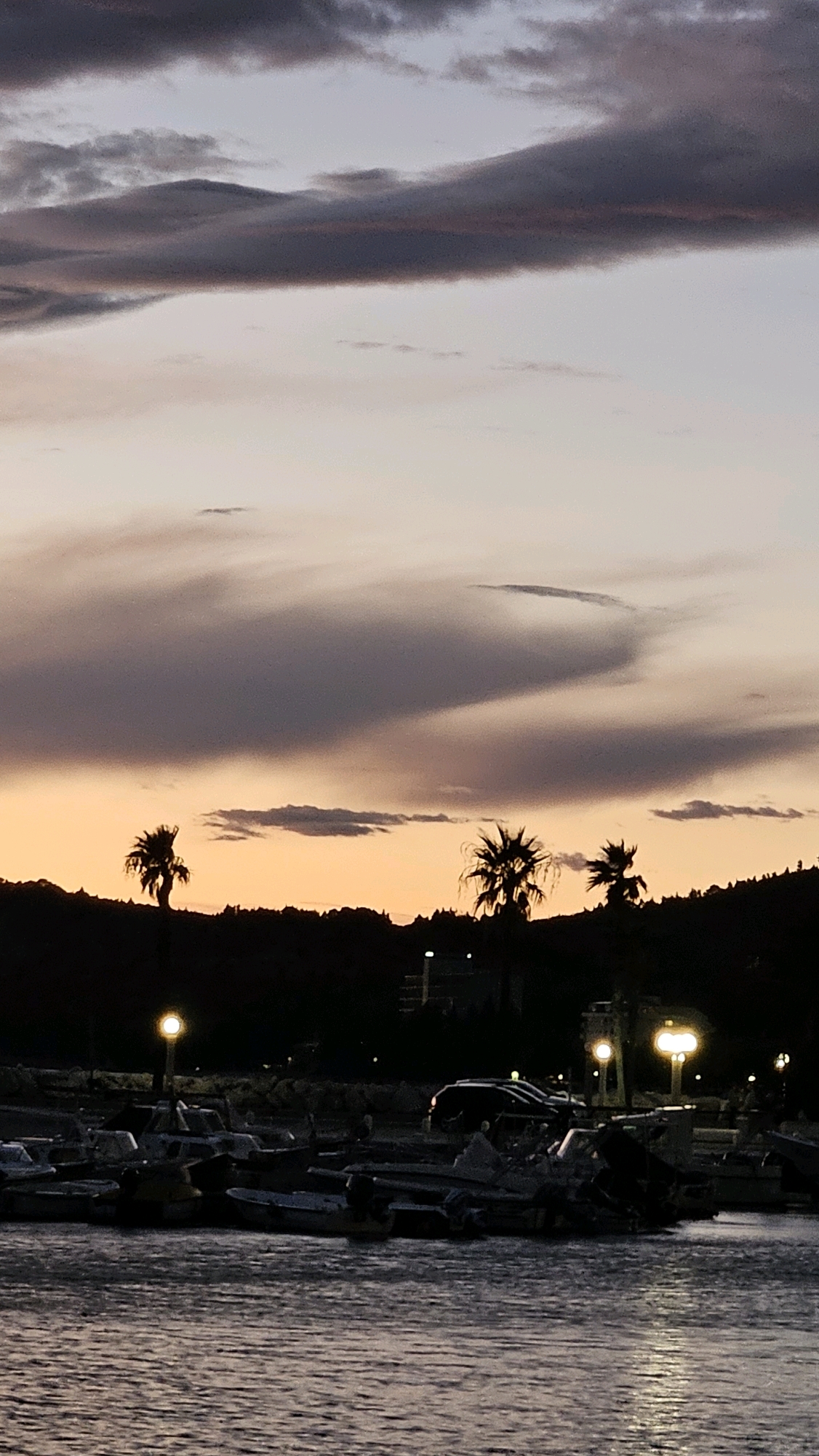 The image captures a twilight scene in Općina Tučepi, Croatia. The sky is filled with dramatic, swirling clouds in shades of gray, purple, and soft orange, suggesting the end of a sunset. Silhouetted against the colorful sky are palm trees and a treeline along a hill. In the foreground, a body of water reflects the dim light, and a harbor is visible with several small boats moored. Lights from streetlamps and possibly buildings are illuminated along the waterfront, casting a warm glow on the scene. The overall atmosphere is tranquil and serene, characteristic of a coastal evening.