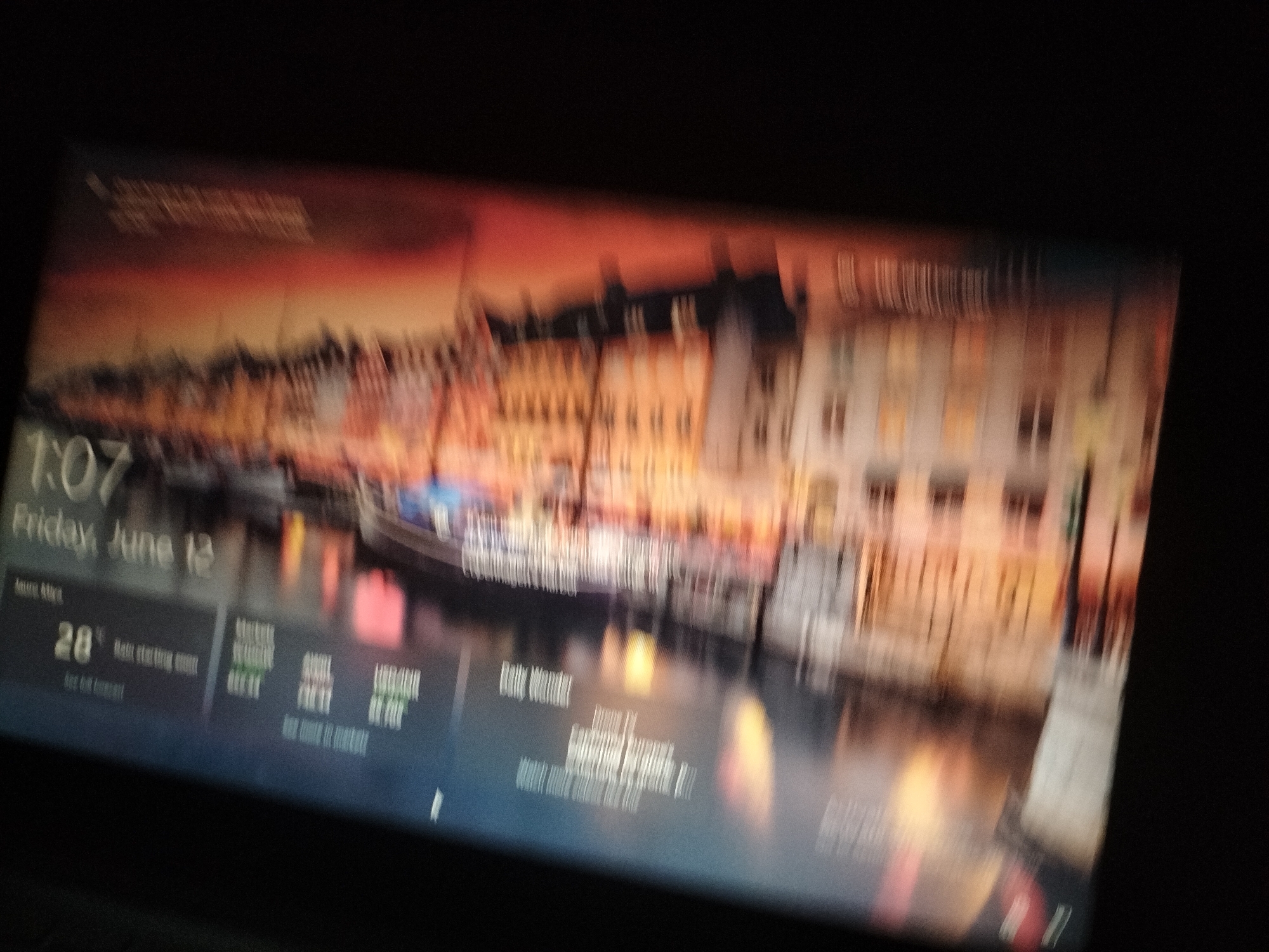 The image displays a digital screen, likely a computer or tablet, featuring a lock screen or desktop wallpaper. The wallpaper depicts a blurred scenic view of buildings along a waterfront, with warm hues of orange and yellow suggesting either sunrise or sunset. Prominently displayed on the screen are the time "1:07" and the date "Friday, June 12". Below this, a weather widget shows a temperature of "28°" and the text "Rain starting soon". Further to the right, other widgets display information related to markets, possibly stock market updates, with abbreviations like "DJIA 10711" and "NASDAQ 4811". The overall scene is one of a digital interface, not a physical location in Jalingo, Nigeria.