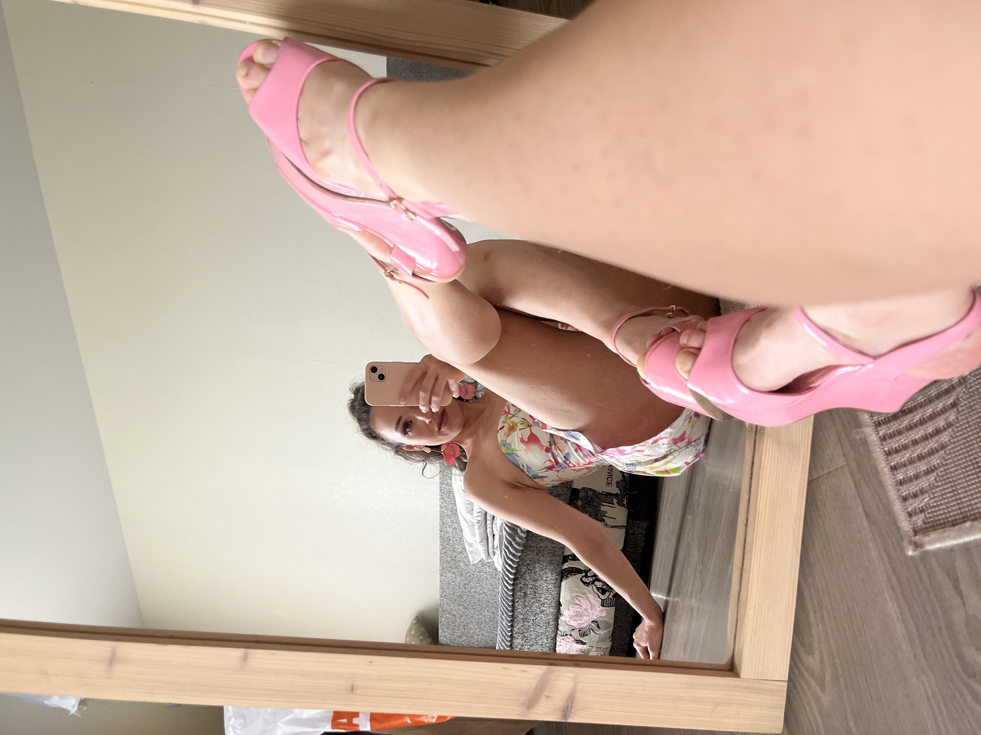 A woman is sitting on the floor in front of a large wooden framed mirror, taking a selfie with her phone. She is wearing a strapless dress with a floral pattern and bright pink wedge sandals. Her left arm is resting on the floor, and her legs are bent. The mirror reflects her entire body, her face, and the room behind her. The room appears to be a living room with a grey sofa and a rug on the wooden floor. The wall behind the sofa is painted a light color. The image does not provide any information about the location being in Kazan, Russia.