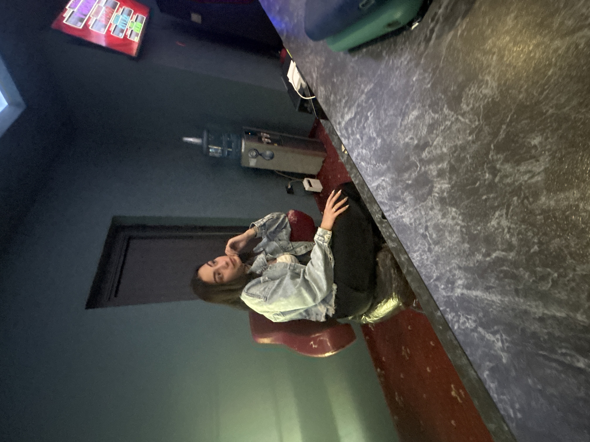 A young woman is seated in a red chair, facing away from the camera and looking towards the left side of the frame. She is wearing a light blue denim jacket over a white top and black pants. Her right hand is resting on her chin, and her left hand is on her lap. The background shows a wall painted in a teal hue, with a dark brown door to the left. To the right of the woman, a water cooler with a blue jug is visible, along with a white electrical outlet and extension cord. In the foreground, a dark gray textured countertop extends across the lower portion of the image, with a green object partially visible on its right side. The scene appears to be indoors, possibly in a bar or lounge area. The image does not provide any specific location details within the city of Beloslav, Bulgaria.
