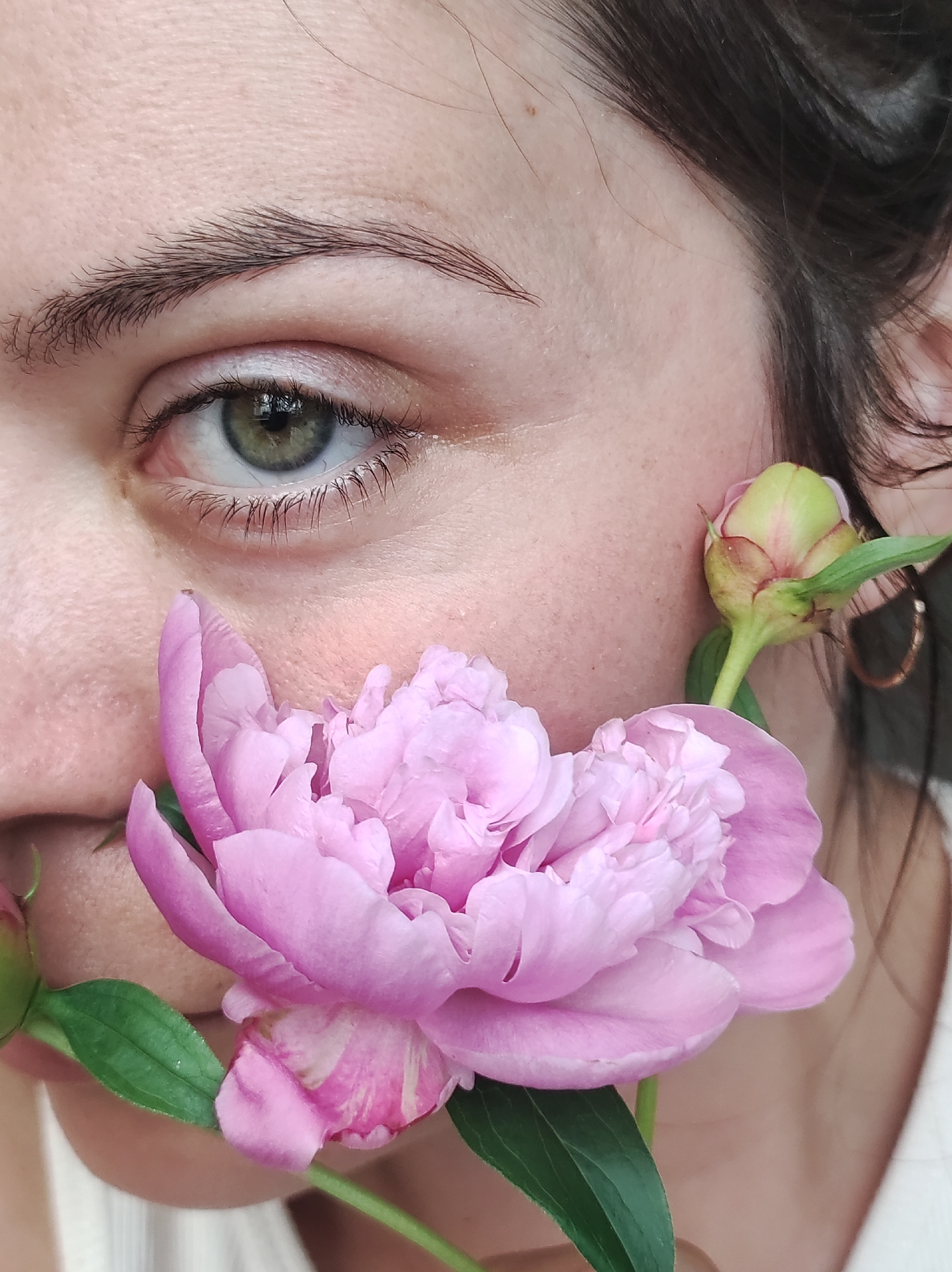 The image is a close-up shot of a person's face, with a pink peony flower partially obscuring their mouth. The person's left eye, with green irises and dark eyelashes, is visible, as is a portion of their eyebrow. The skin on the cheek shows some texture and a slight blush. A closed peony bud is visible near the ear, and a gold hoop earring can be seen in the right ear. Dark hair frames the right side of the face. The background is blurred and indistinct, suggesting an outdoor setting. There is no discernible information within the image that would indicate its location in Chaikovsky, Russia.