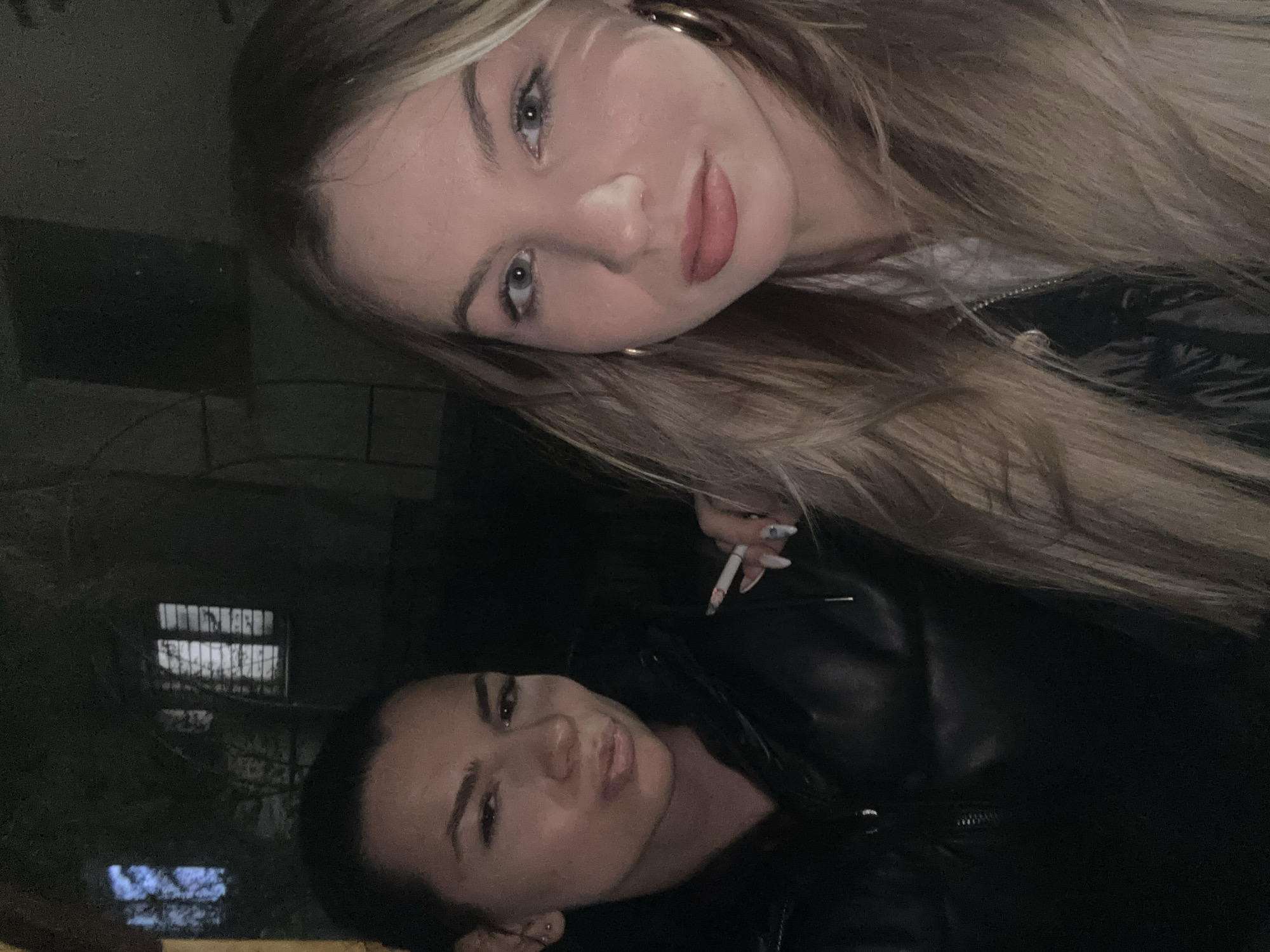 Two women are photographed at night. The blonde woman has long hair, gold hoop earrings, and red lipstick. The other woman has dark hair and is wearing a black leather jacket. The blonde woman holds a lit cigarette. In the background, there is a building with barred windows. The location is in Plovdiv, Bulgaria.