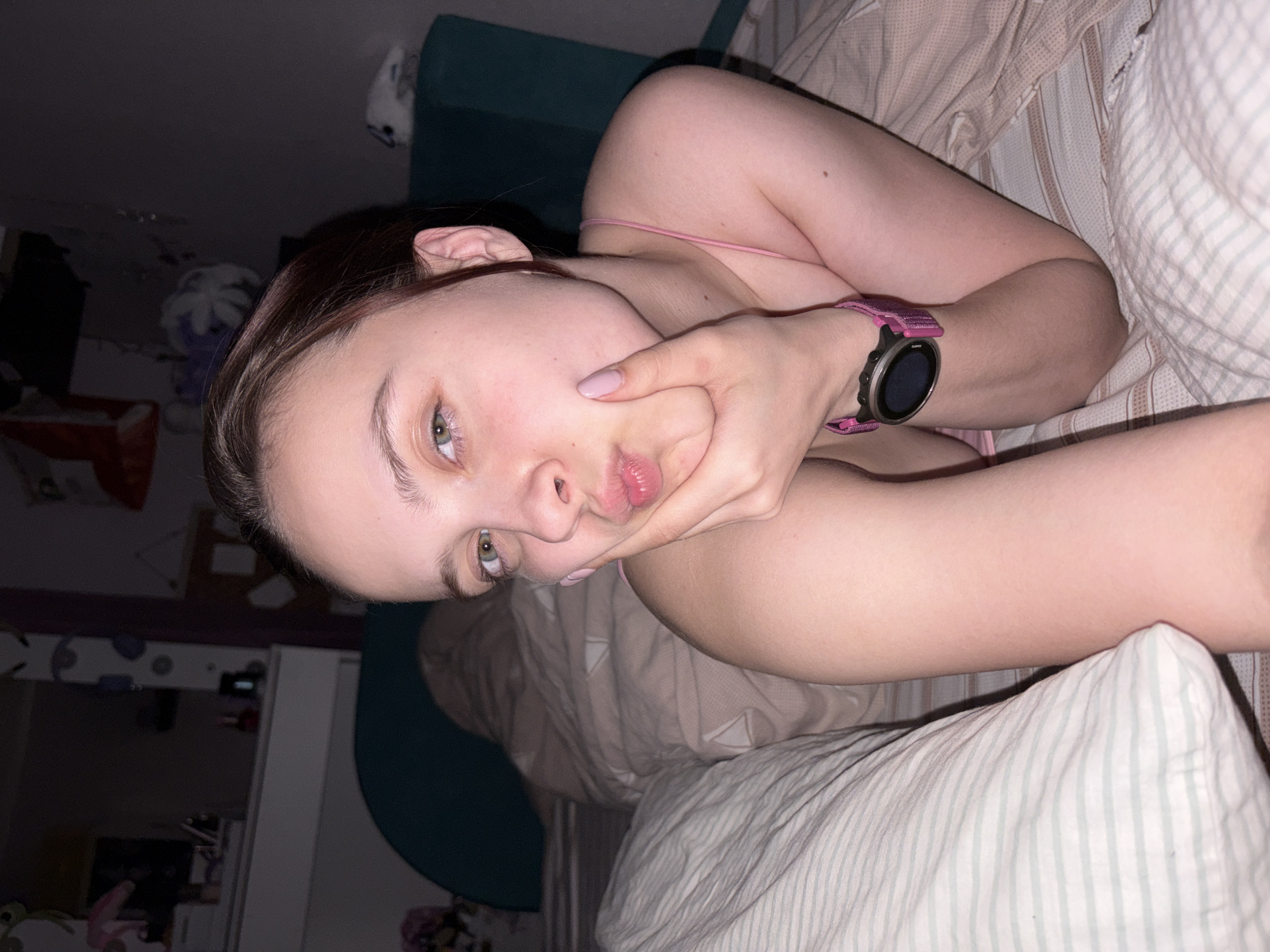 The image, rotated counter-clockwise, displays a close-up of a young person lying down indoors, likely in a bedroom, under low light conditions. The main subject is a light-skinned person with light brown hair pulled back, revealing their face. They have striking greenish-hazel eyes and a light complexion.
The person is looking directly at the camera with a slightly pouting expression, their left hand (with light pink nail polish) gently pressed against their cheek and chin, accentuating the pout. They are wearing a pink spaghetti strap top. On their left wrist, a digital sports watch with a black circular face and a vibrant pink fabric strap is clearly visible.
The background suggests an intimate indoor setting, likely a bed. Parts of striped bedding, including pillows and a duvet, are visible in shades of white and light brown/beige. Behind the subject's head, a dark teal or green upholstered headboard or cushion can be seen. Further back, the room is dimly lit, showing hints of furniture, shelves, and general room clutter, indicating it's likely nighttime or very early morning. The overall mood is casual and personal, characteristic of a selfie. No visible text is present in the image. The location context provided is Прикамский, Russia.