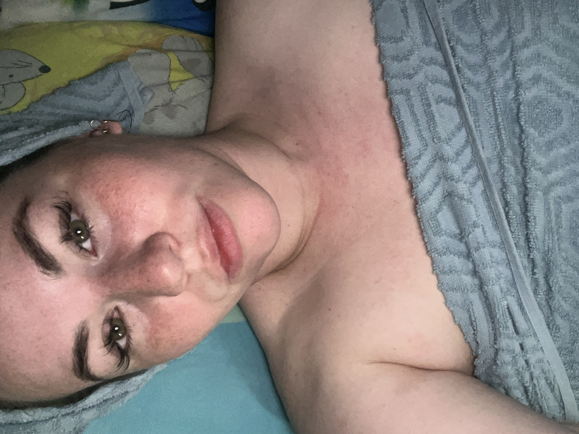 A close-up, slightly rotated portrait captures a fair-skinned individual lying down indoors, likely in a bedroom in Chaikovsky, Russia. The subject, who appears to be a female, looks directly at the camera with a relaxed expression. Her face shows freckles and some redness on her skin and décolletage. She has dark, distinct eyebrows, long dark eyelashes, and green or hazel eyes. Her lips are a soft pinkish-red.
A light blue, patterned towel is visible, partially wrapped around her head and covering her upper body. A small, silver hoop earring adorns her visible ear. She is resting on a bed with bedding consisting of a light blue sheet or pillowcase and a second pillow featuring a yellow and blue pattern with grey, cartoon-like animal designs. The lighting is dim, suggesting an evening or nighttime setting, with hints of artificial light reflecting in her eyes. No text is visible in the image.