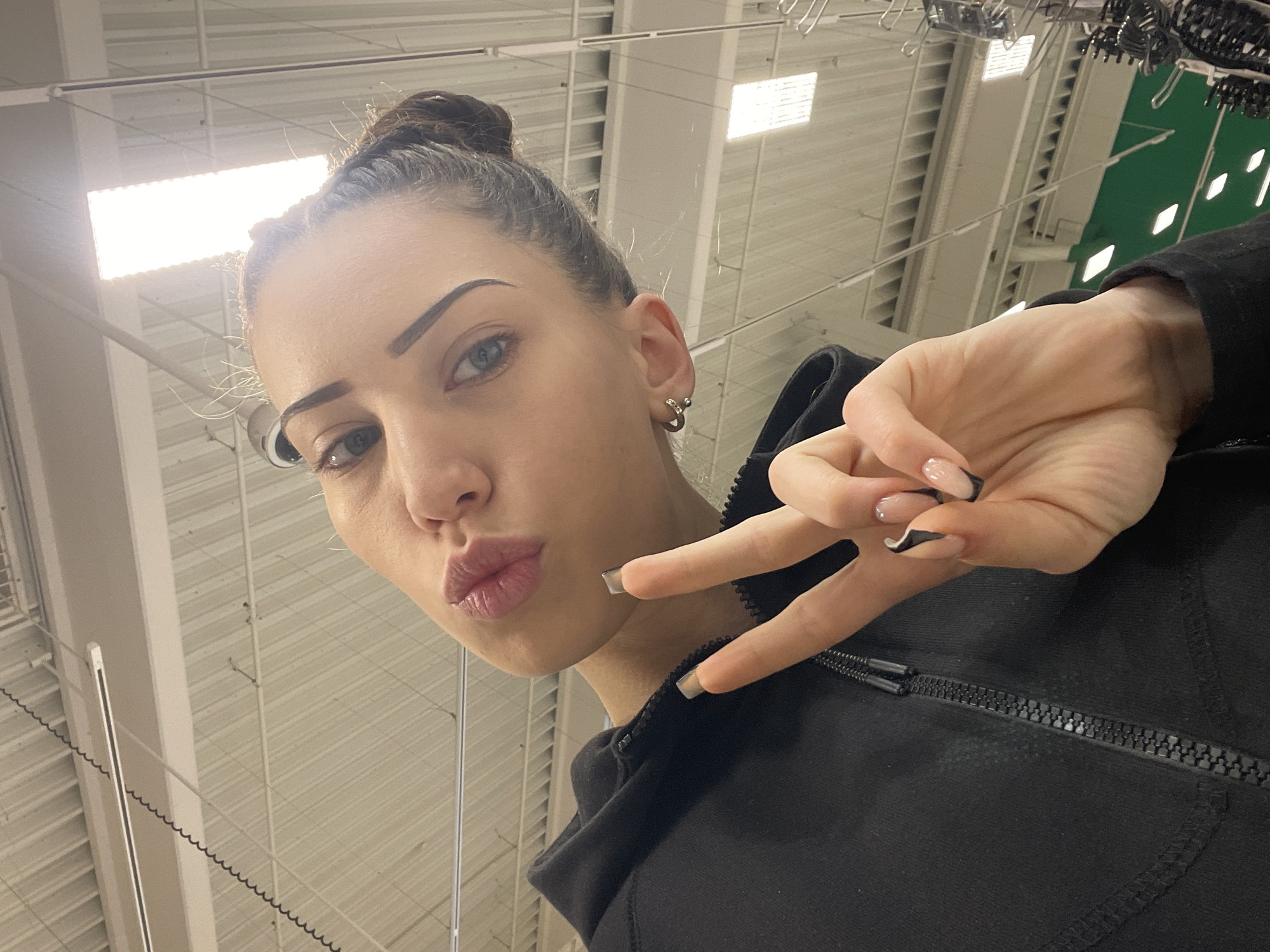 A low-angle selfie shows a woman with her hair in a bun, making a peace sign with her right hand. She has fair skin, prominent cheekbones, and dark eyebrows. She is wearing a black zippered hoodie. The background consists of an industrial-style ceiling with fluorescent lighting and some kind of ventilation system. The location is inside a commercial building; however, based on the information provided, it is impossible to determine if the image was taken in the city of Veliko Tarnovo, Bulgaria.
