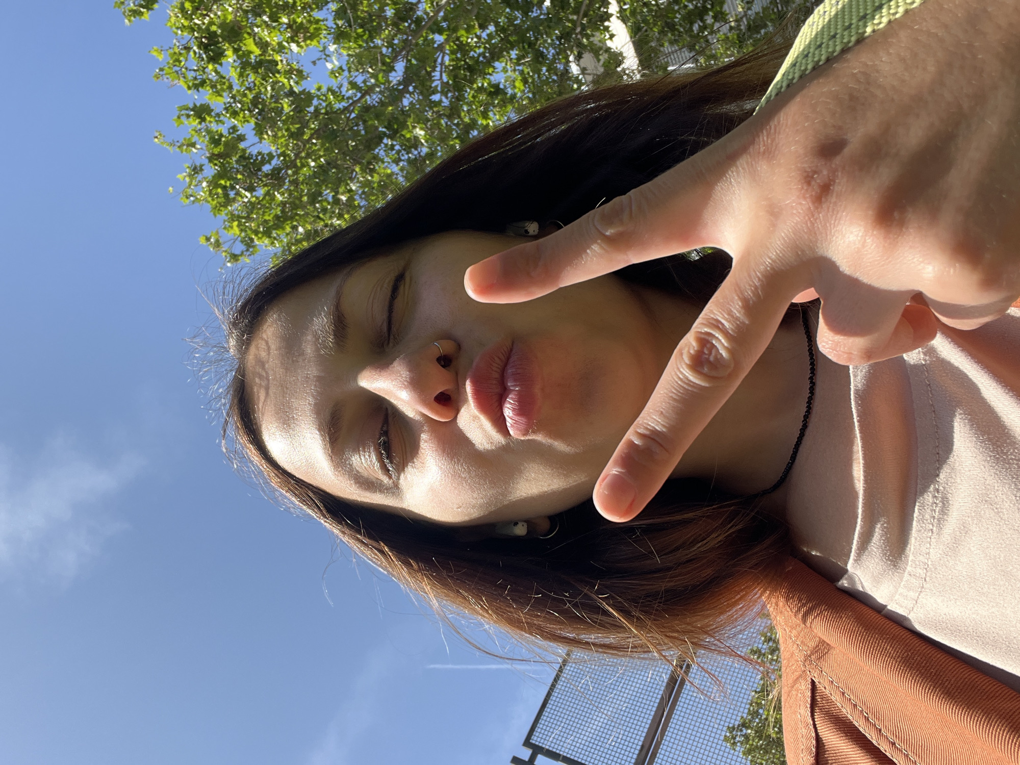 A woman is taking a selfie against a backdrop of a bright blue sky with a few wispy clouds, and green foliage in the upper left. Her dark hair is parted and falls around her face. She is wearing earbuds and has a nose ring. Her lips are pursed. One hand is raised, forming a peace sign. She is wearing a light-colored t-shirt with a brown jacket. Part of a chain-link fence is visible in the lower left. Based on the trees and fence, it is possible the location is in Barcelona.