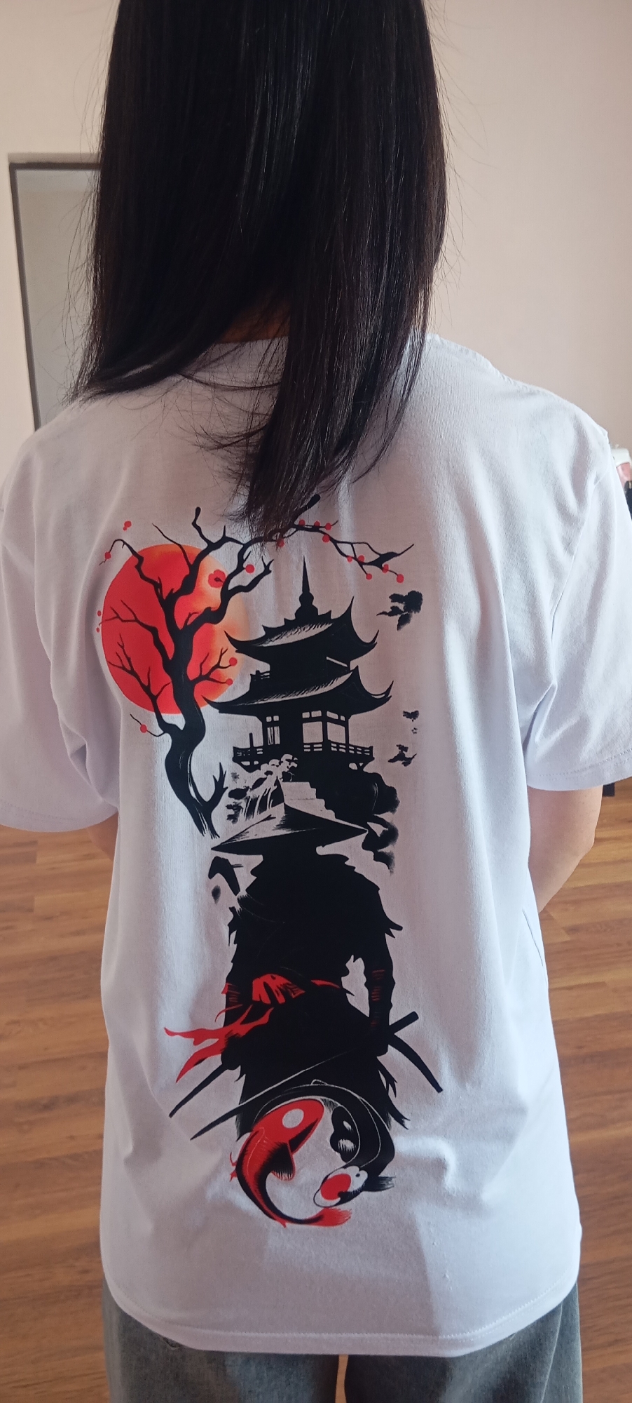 The back of a white t-shirt is displayed, featuring a vertical graphic with Japanese-inspired imagery. The design includes a silhouette of a pagoda, a leafless tree with a red sun behind it, a samurai warrior in silhouette with a straw hat, and two koi fish in a yin-yang formation at the bottom. The t-shirt is worn by a person whose back and dark hair are visible, standing in an indoor environment with wooden flooring and a light-colored wall in the background. The image is a medium shot, focusing on the t-shirt's graphic. There is no indication of the city of Baksan, Russia in this image.