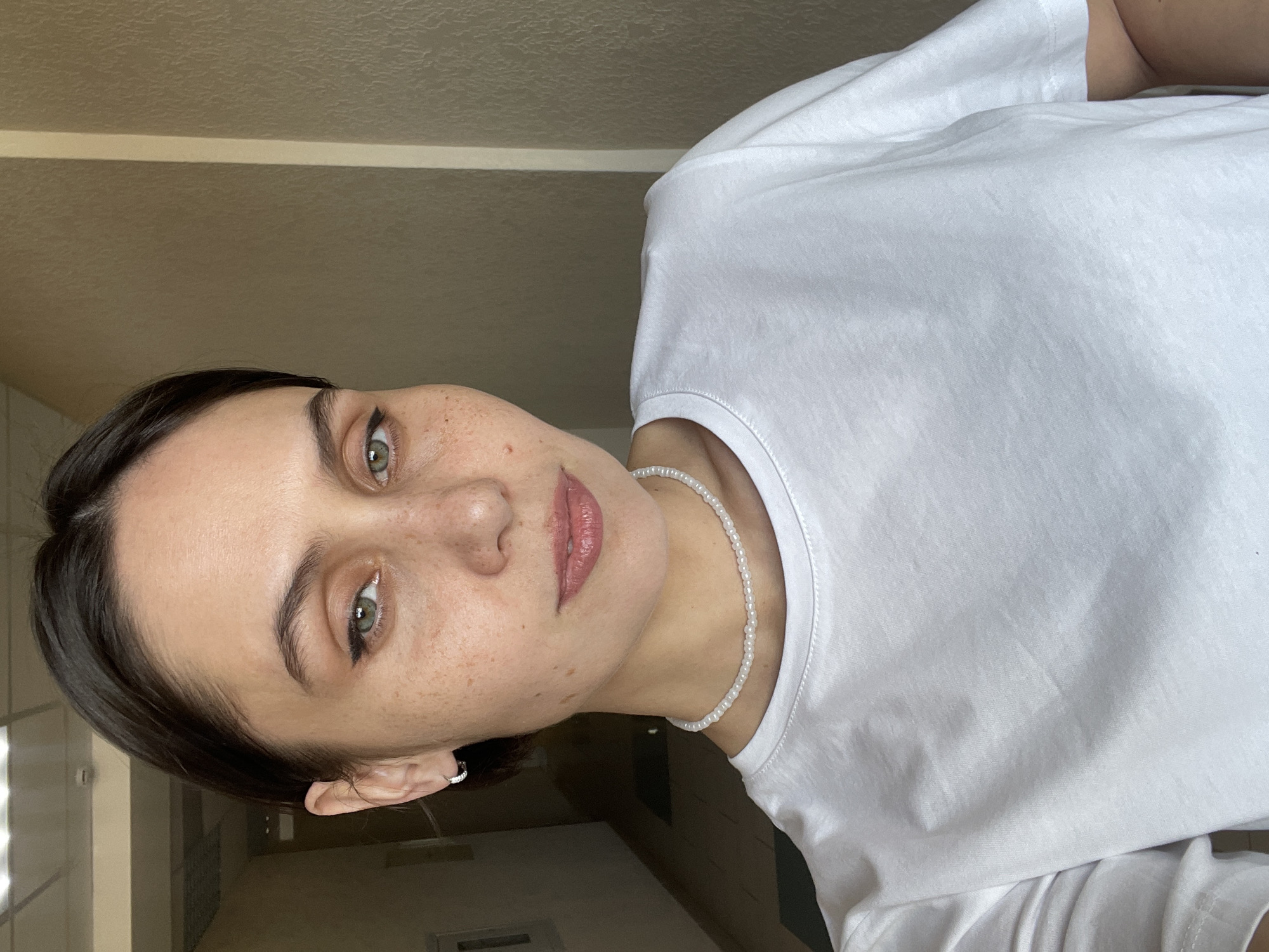A woman with fair skin and dark hair poses for a selfie indoors. She wears a white t-shirt and a white beaded necklace. Her left ear features a small hoop earring. Freckles are visible on her face. The background is a beige wall and hallway. It is not possible to determine the location as Izhevsk, Russia from the given image.