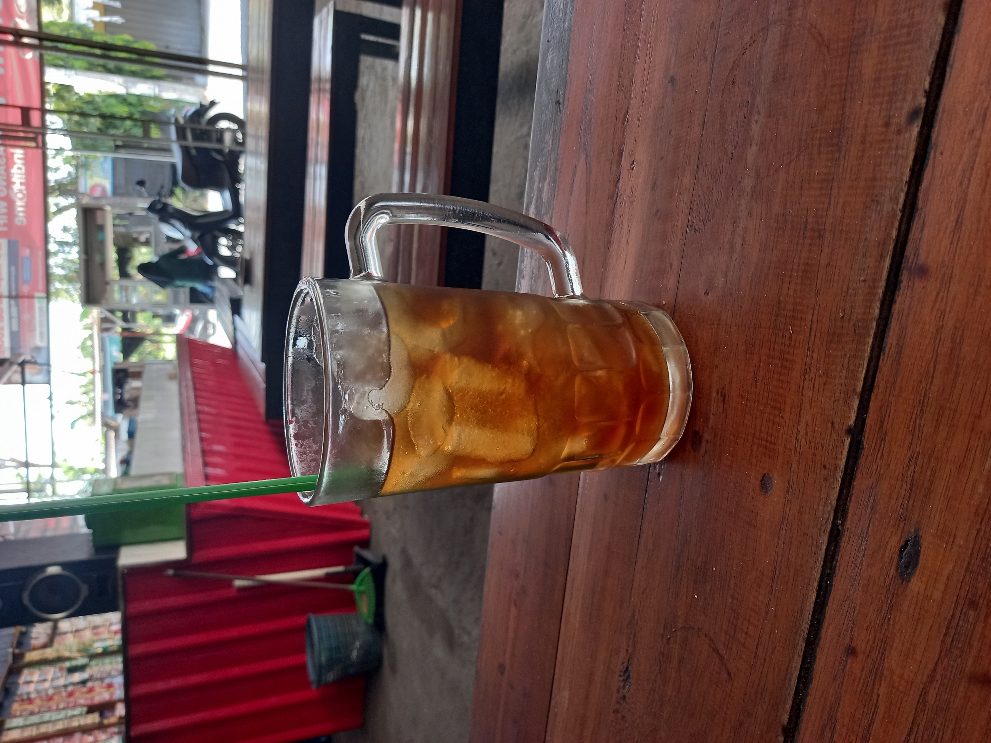 A close-up shot captures a tall glass of iced tea, filled with ice cubes and a green straw, resting on a dark wooden table. The glass is positioned in the foreground, slightly off-center to the right. Behind the table, a blurred scene unfolds, suggesting an outdoor seating area of a roadside food stall or cafe. Red corrugated metal barriers are visible to the left, and in the background, a motorcycle is parked near a food cart. A person wearing a hijab is visible in the background near the food cart, potentially a vendor or customer. The overall setting appears to be an open-air establishment in Pelemwatu, Indonesia, likely a casual dining or rest stop.