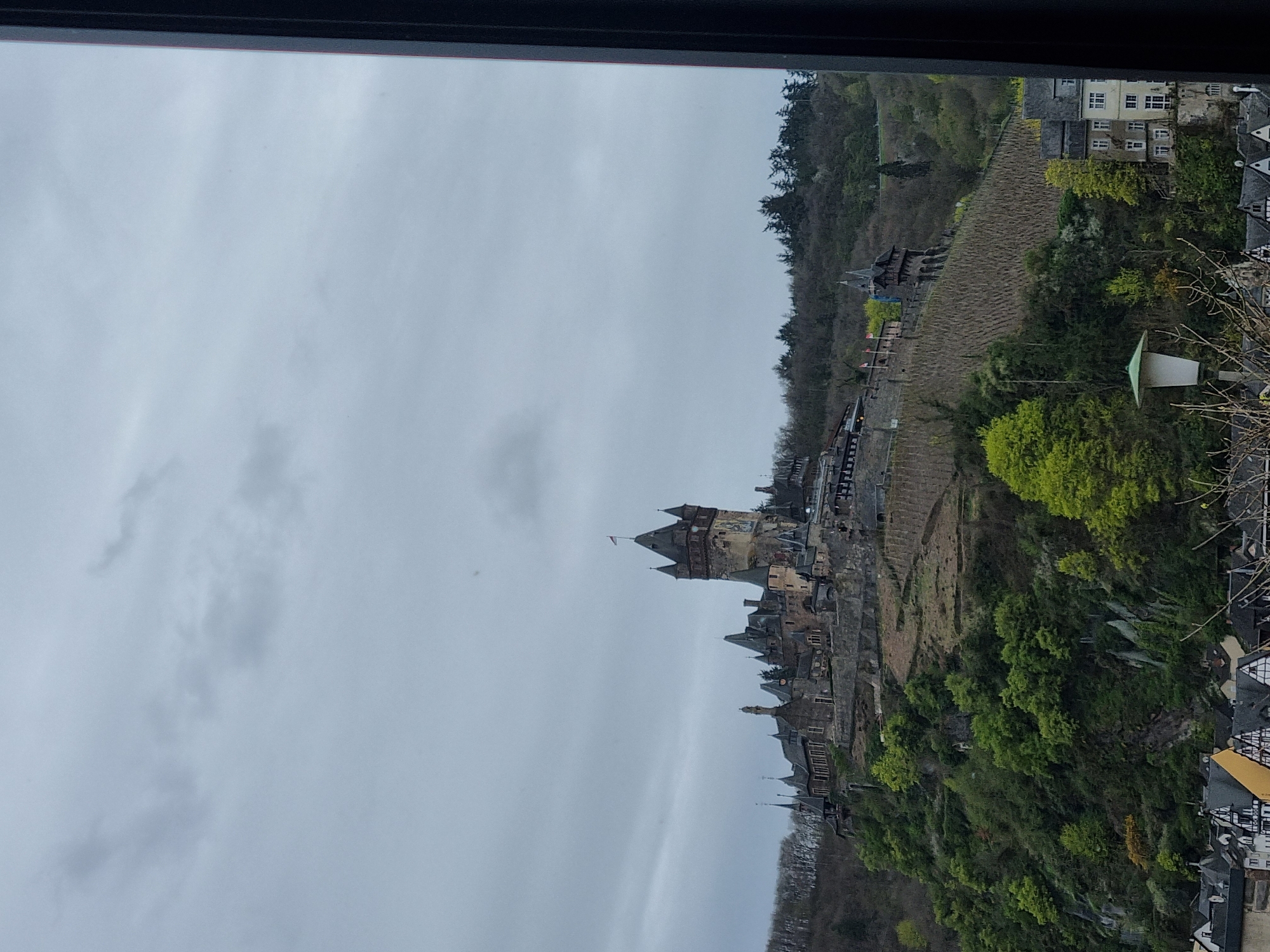 The image captures a view from a window, looking out onto the town of Cochem, Germany, dominated by the Reichsburg Cochem castle. The castle is built on a hill overlooking the town. Vineyards cover the slopes of the surrounding hills. The sky is overcast and grey. Buildings line the riverbank.