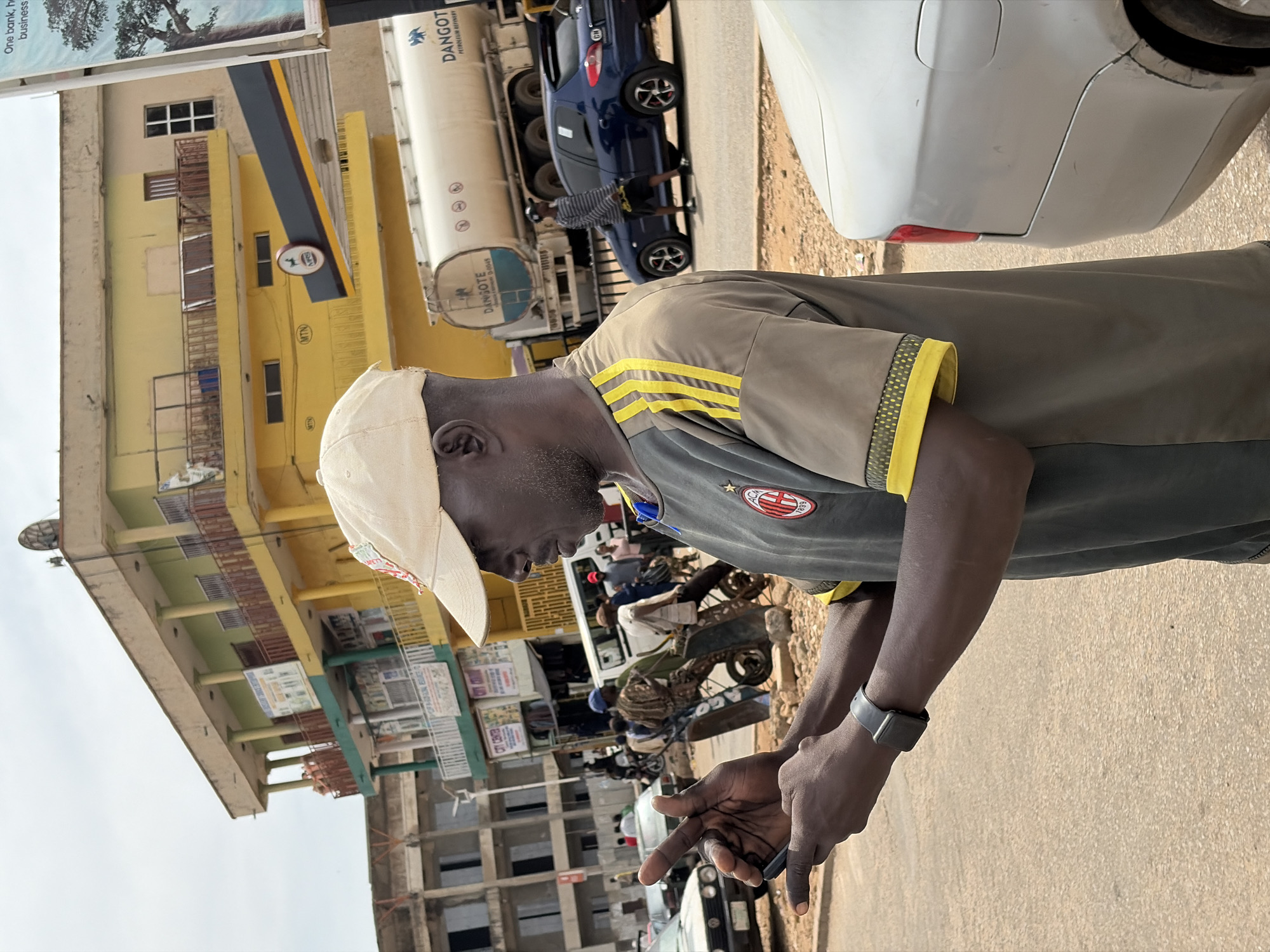 A man in a tan cap and a grey and yellow t-shirt holds a smartphone in his hands, appearing to text or browse. He is standing on a street in Jos, Nigeria, with buildings and vehicles in the background. To his left, there are cars and construction sites. To his right, a white Dangote petroleum tanker truck and a blue car are visible. A man in a striped shirt walks by the blue car. The scene is likely a busy street with commercial activity.