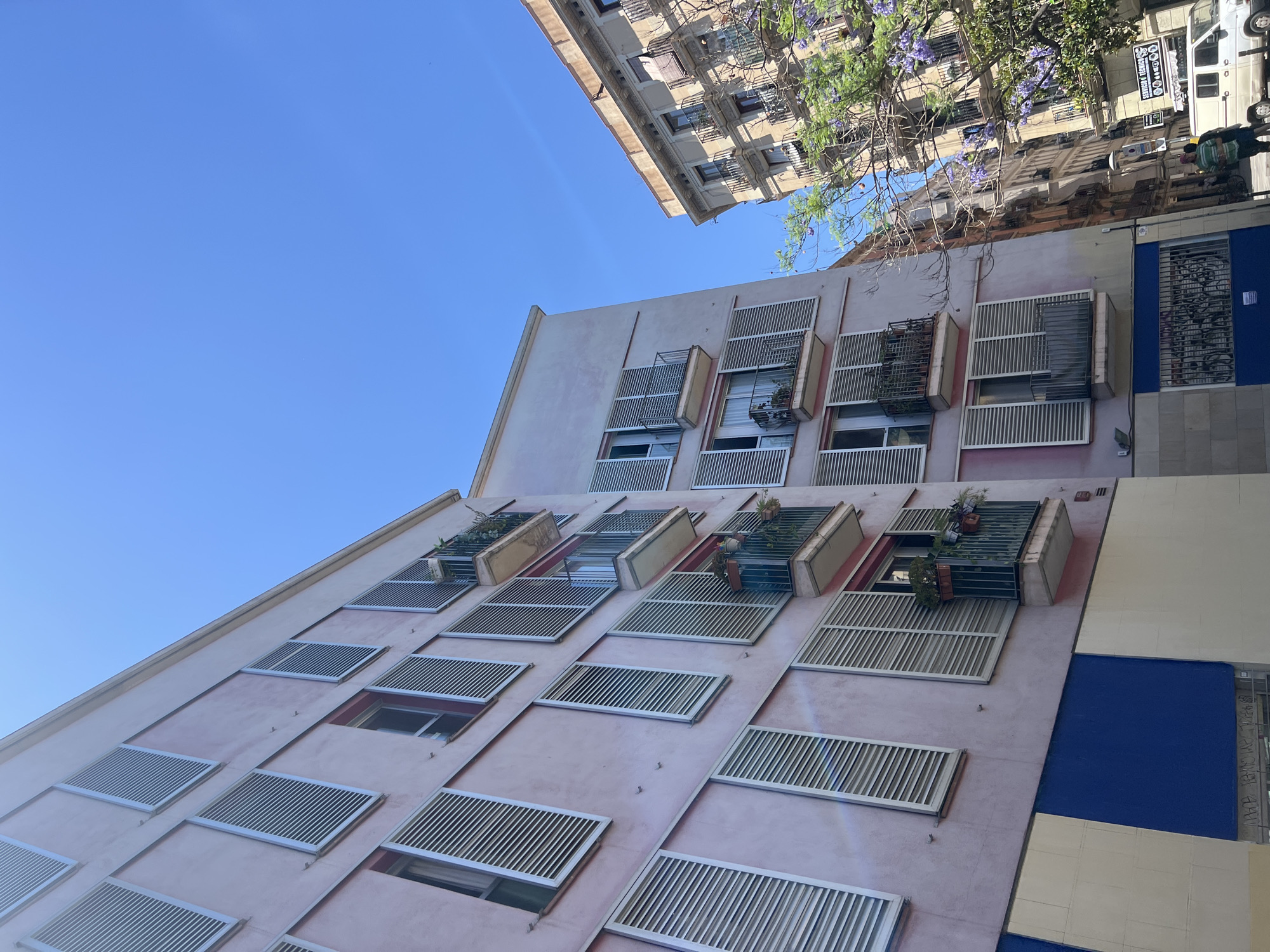A low-angle, slightly tilted shot captures the facades of two apartment buildings against a clear blue sky in Barcelona, Spain. The building on the left, painted a pale pink, features multiple stories with uniformly shuttered windows. Some balconies jut out, adorned with potted plants. The lower portion of this building has a contrasting blue and cream tiled facade. To the right, a taller, older building with ornate balconies and a beige facade stands, partially obscured by the branches of a tree with purple blossoms. A white van and two people are visible on the street below the buildings. The scene depicts a typical urban streetscape with residential architecture.