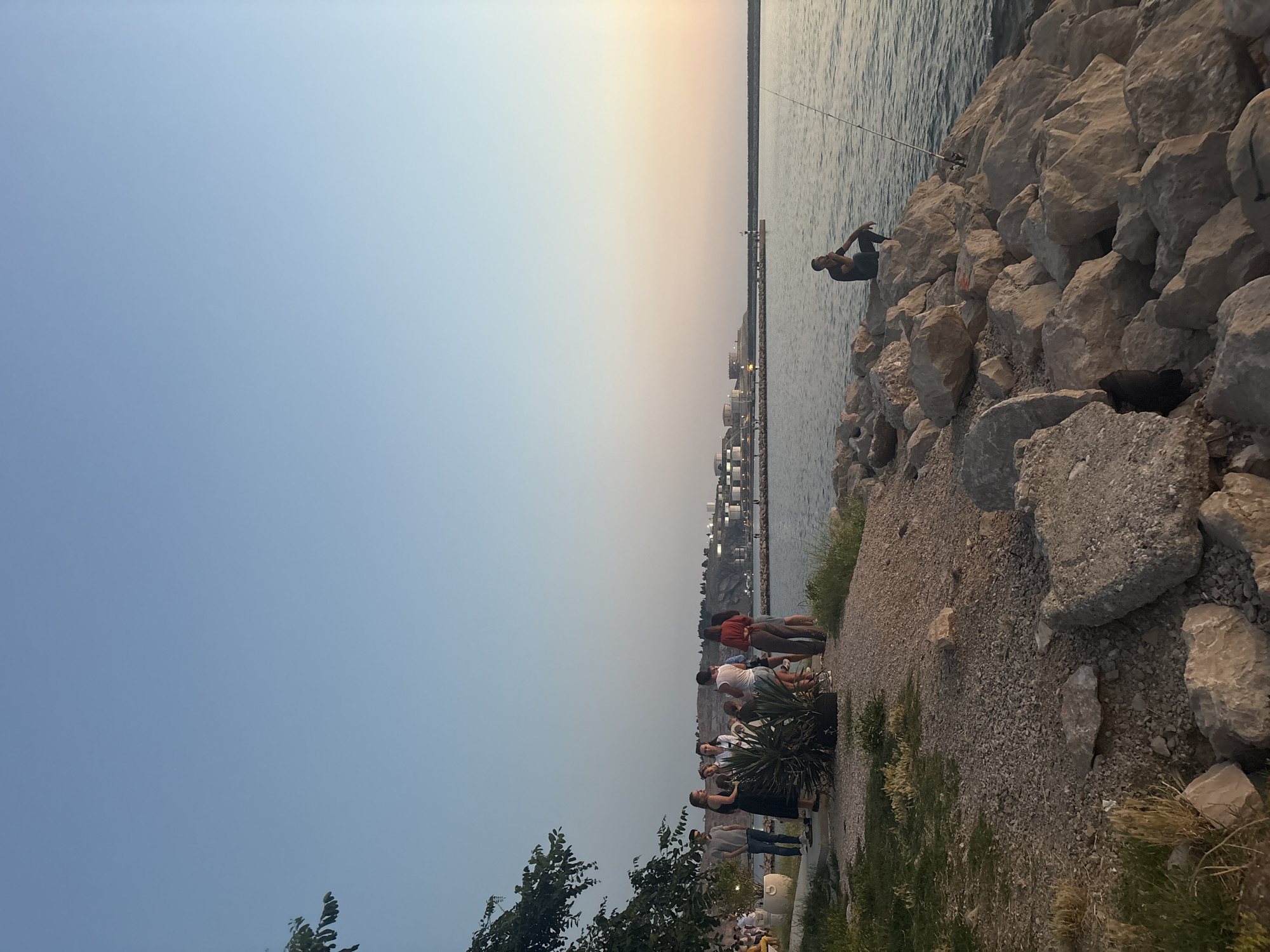 The image captures a serene evening scene by the sea in Bar, Montenegro. A group of people are gathered on a pebble-strewn embankment on the left side of the frame, some standing and conversing, while others are seated. A man is seen fishing on a rocky outcrop to the right, with his fishing rod extended into the calm water. In the background, a harbor with several buildings and a lighthouse is visible across the bay. The sky transitions from a clear blue overhead to a soft gradient of orange and pink near the horizon, indicating the approach of sunset. Trees and vegetation are present along the shoreline, adding to the natural beauty of the location.