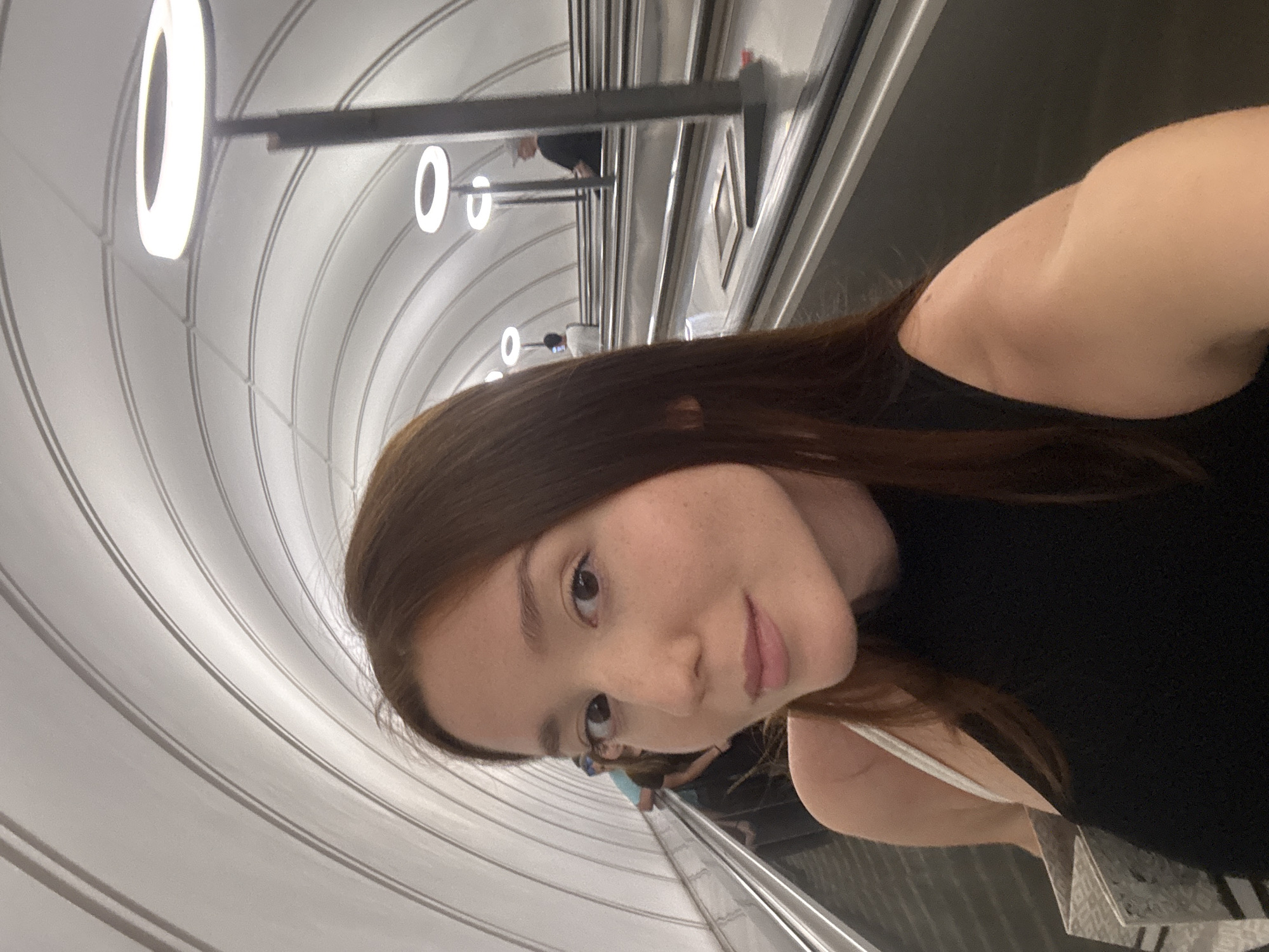 A woman with long brown hair takes a selfie on a modern escalator in what appears to be a subway station in Khimki, Russia. She is wearing a black sleeveless top and a white strap is visible over her left shoulder. She is holding a patterned shopping bag. The escalator is flanked by metallic railings and the ceiling of the station features a distinctive arched design with evenly spaced ring-shaped lights. In the background, other individuals can be seen on parallel escalators. The scene suggests a functional and contemporary urban transit environment.