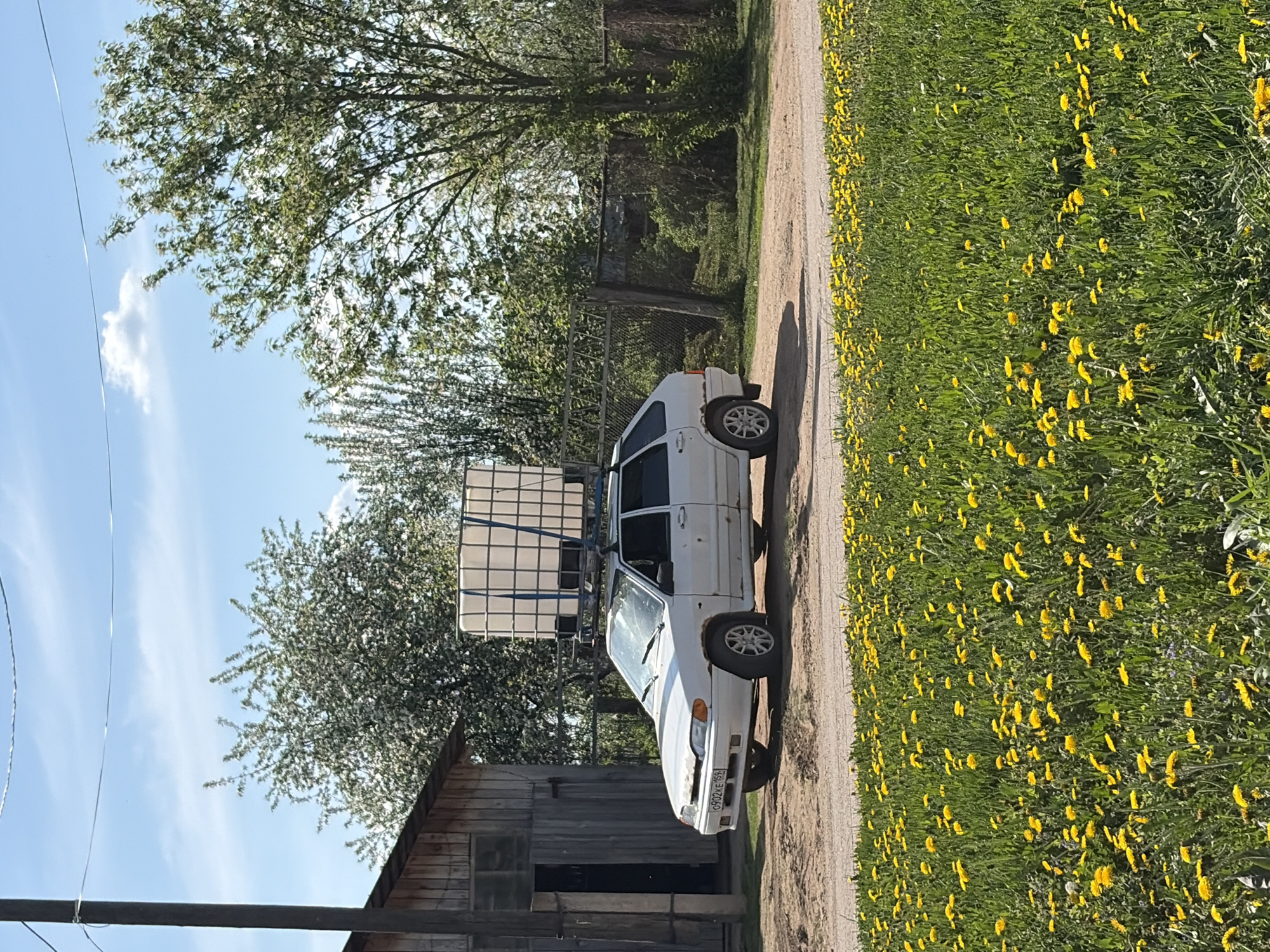 The image depicts a white car with a large rectangular container secured to its roof rack. It is parked on a dirt road adjacent to a field of yellow wildflowers. A weathered wooden structure is visible to the left of the car. The background includes trees and a fence. The sky is blue with a few clouds. The license plate reads C902KE 53. The location is assumed to be Чумна, Russia, based on the request.