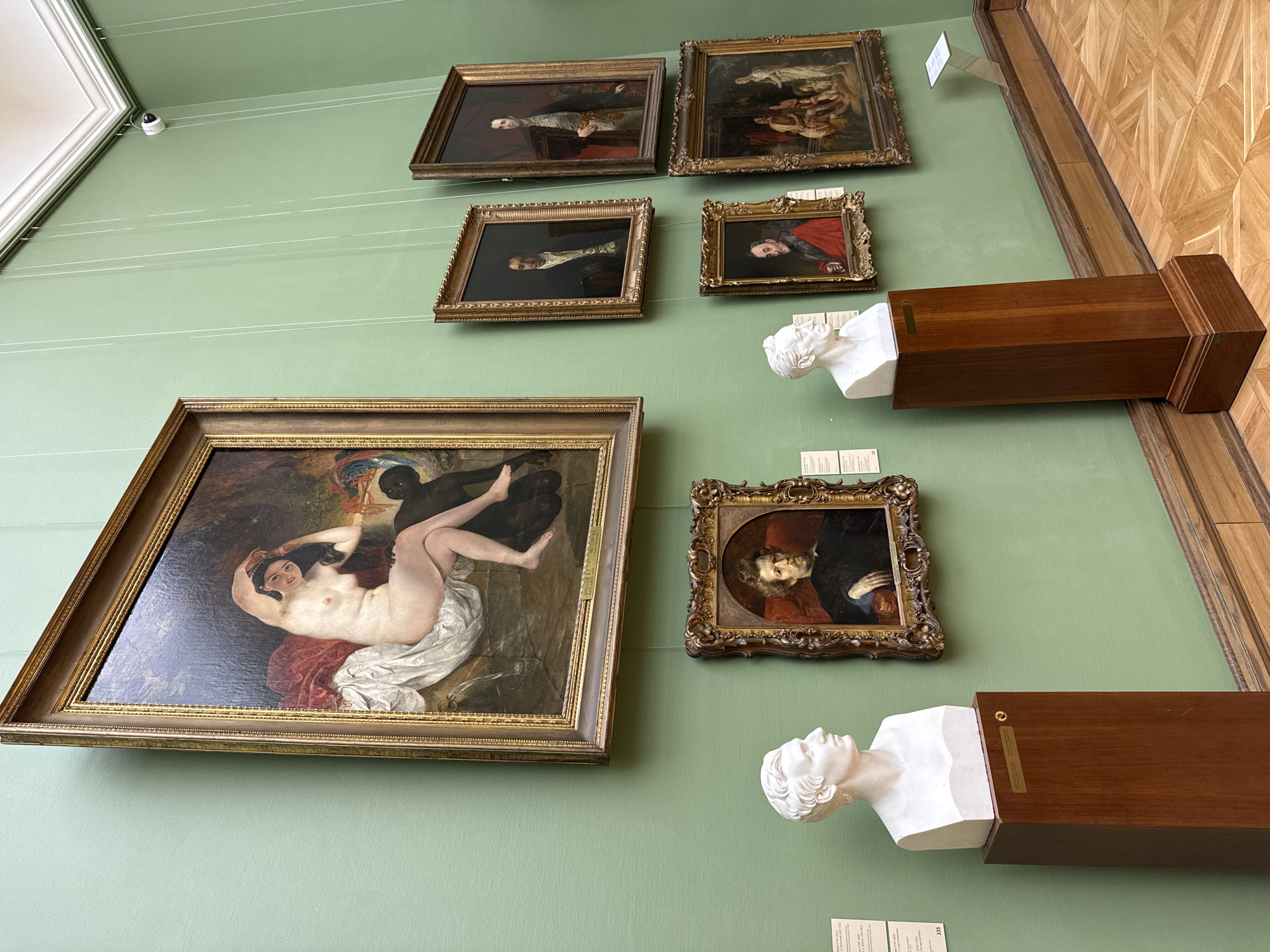 The image depicts a section of an art gallery, specifically within a location in Moscow, Russia. The scene focuses on a pale green wall adorned with multiple framed paintings and two sculpted busts mounted on wooden plinths.
The paintings, varying in size and frame style, portray figures in classical or historical settings. The largest painting is on the lower left side, depicting what appears to be a nude woman and a black male figure. The other paintings appear to be portraits.
Two white busts are mounted, appearing to be classical figures, on wooden plinths extending from the wall. White labels are below the paintings and busts, providing information about the artworks. The gallery wall is lined with a wooden floor.
A security camera is visible near the top of the wall.