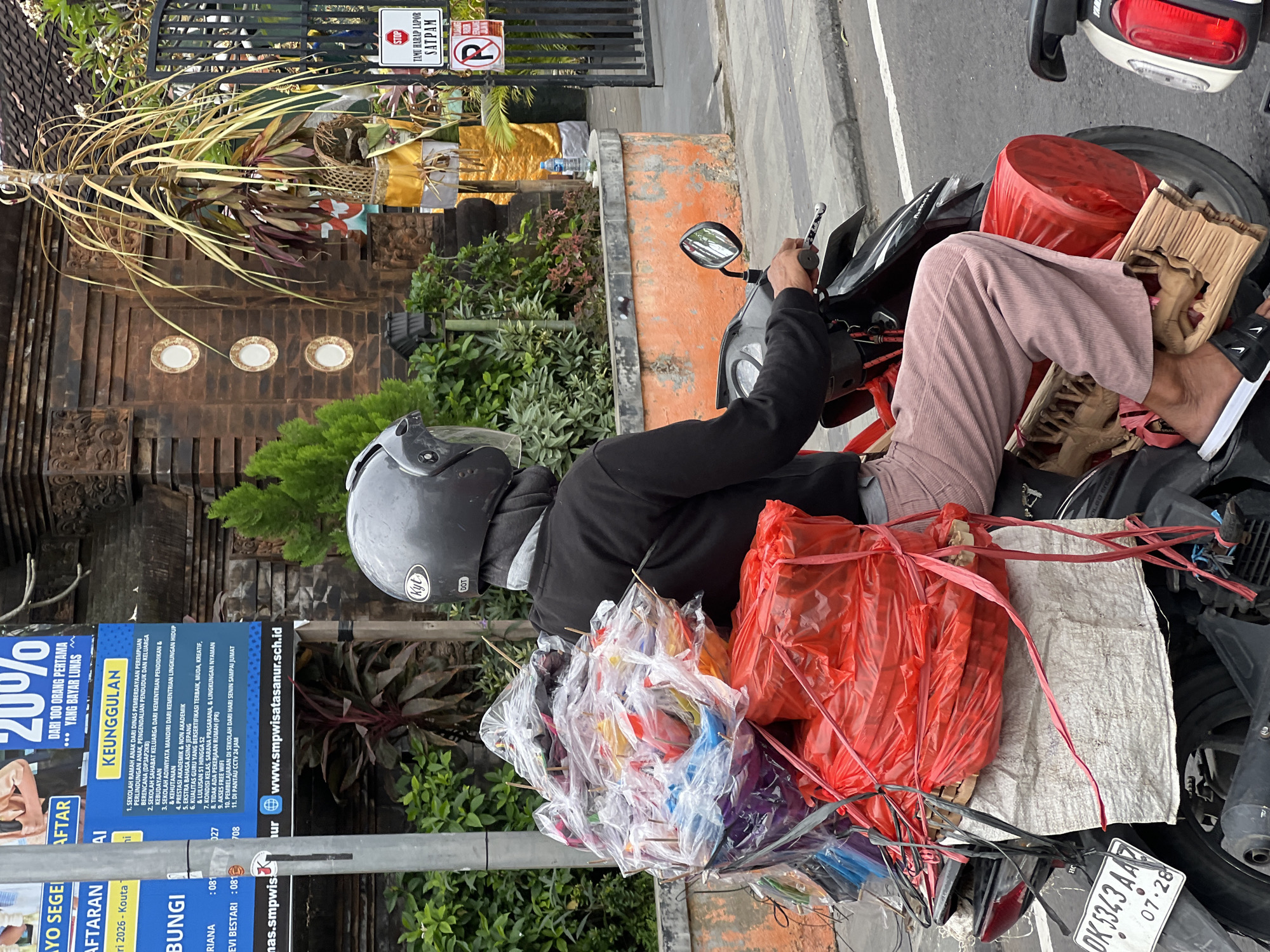 A person wearing a helmet and black long-sleeved shirt rides a motorcycle laden with goods. The motorcycle is black and red, with the license plate "DK 5343 AAZ". The goods are wrapped in various plastic bags and tied with red and black ropes. The person is wearing pink corduroy pants and black and white slides. The background shows a building with intricate carvings, greenery, and a black gate. There is also a stop sign and a parking sign visible. The scene appears to be on a street in Denpasar, Indonesia, given the license plate format and the architectural style.