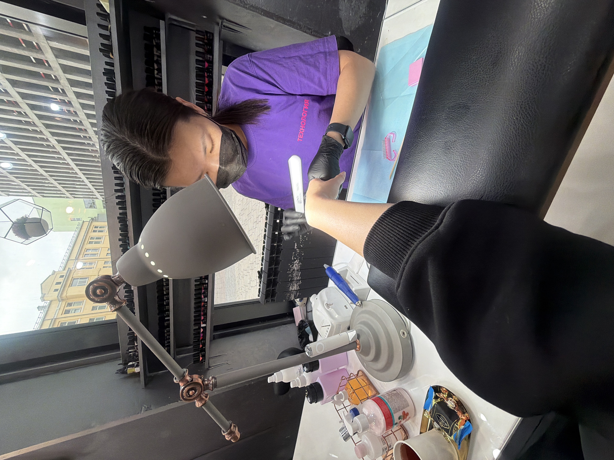 A woman wearing a purple T-shirt and a black face mask is giving a manicure. She is wearing black gloves and holding a white nail file. Her right wrist has a black smartwatch. The person receiving the manicure is wearing a black hoodie. They are sitting at a table in front of the woman. The table has a blue pad and various manicure supplies. A gray lamp stands next to the table. The background shows a building through a window. Based on available details, the location is likely in Khimki, Russia.