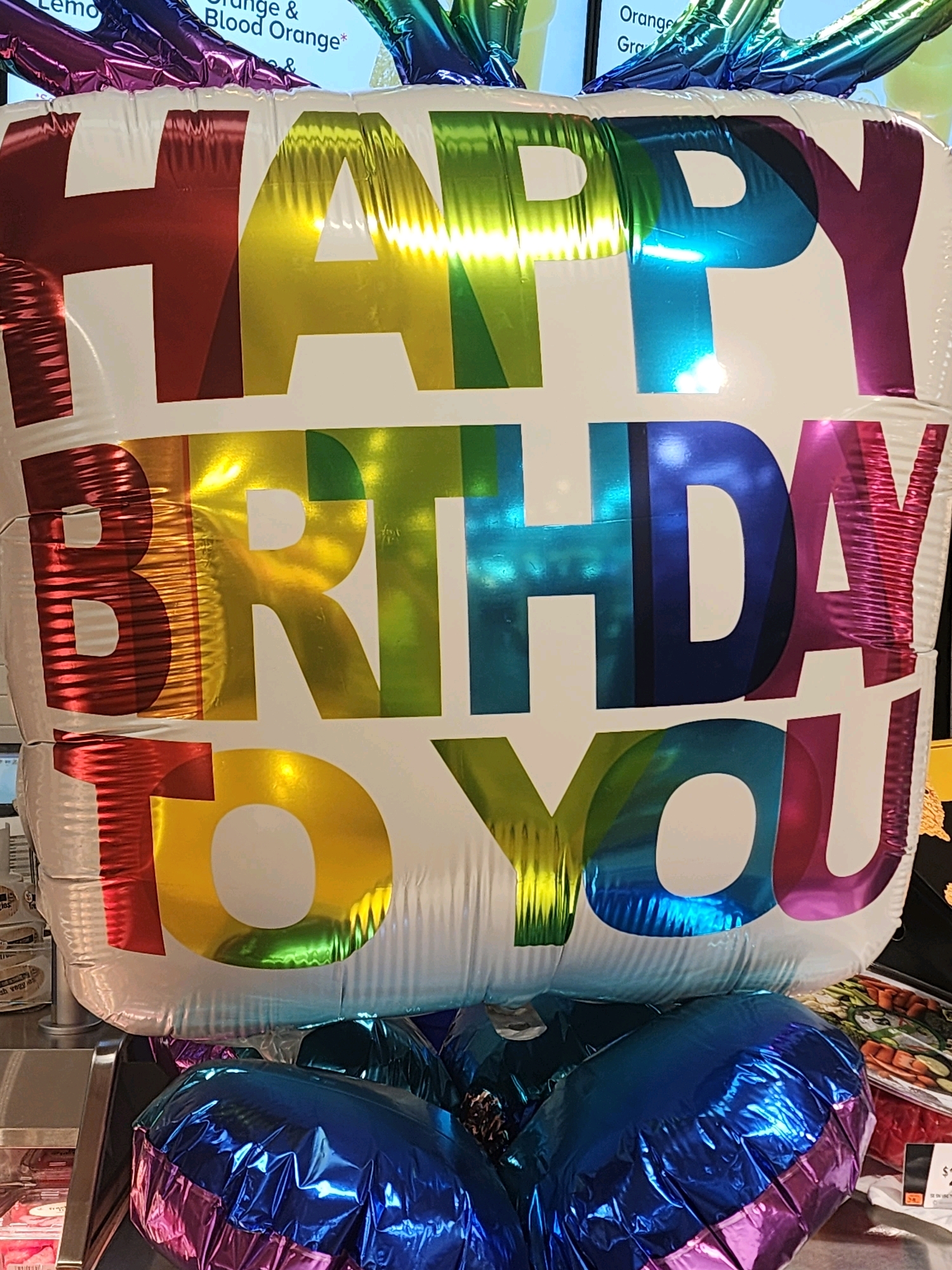 A large, white, Mylar balloon with iridescent rainbow lettering reads "HAPPY BIRTHDAY TO YOU." The balloon is inflated and slightly wrinkled, with a metallic sheen that reflects light in various colors. It is positioned in the foreground, obscuring the lower portion of the image. The balloon appears to be part of a larger balloon arrangement, as evidenced by the partially visible blue and pink balloons beneath it. In the background, a blurry display of products can be seen, suggesting a retail environment. The lighting is bright and somewhat harsh, creating strong highlights and shadows on the balloon.