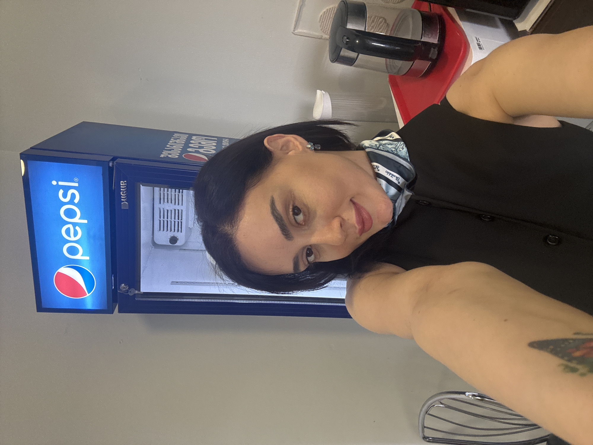 A woman with short, dark hair is pictured in what appears to be an indoor setting. She is wearing a black button-down shirt and a patterned scarf. To her left is a blue Pepsi refrigerator unit. Behind her, there is a kettle on a red tray, indicating a potential office or breakroom setting. The location is potentially in Tbilisi, Georgia, given the branding on the refrigerator.