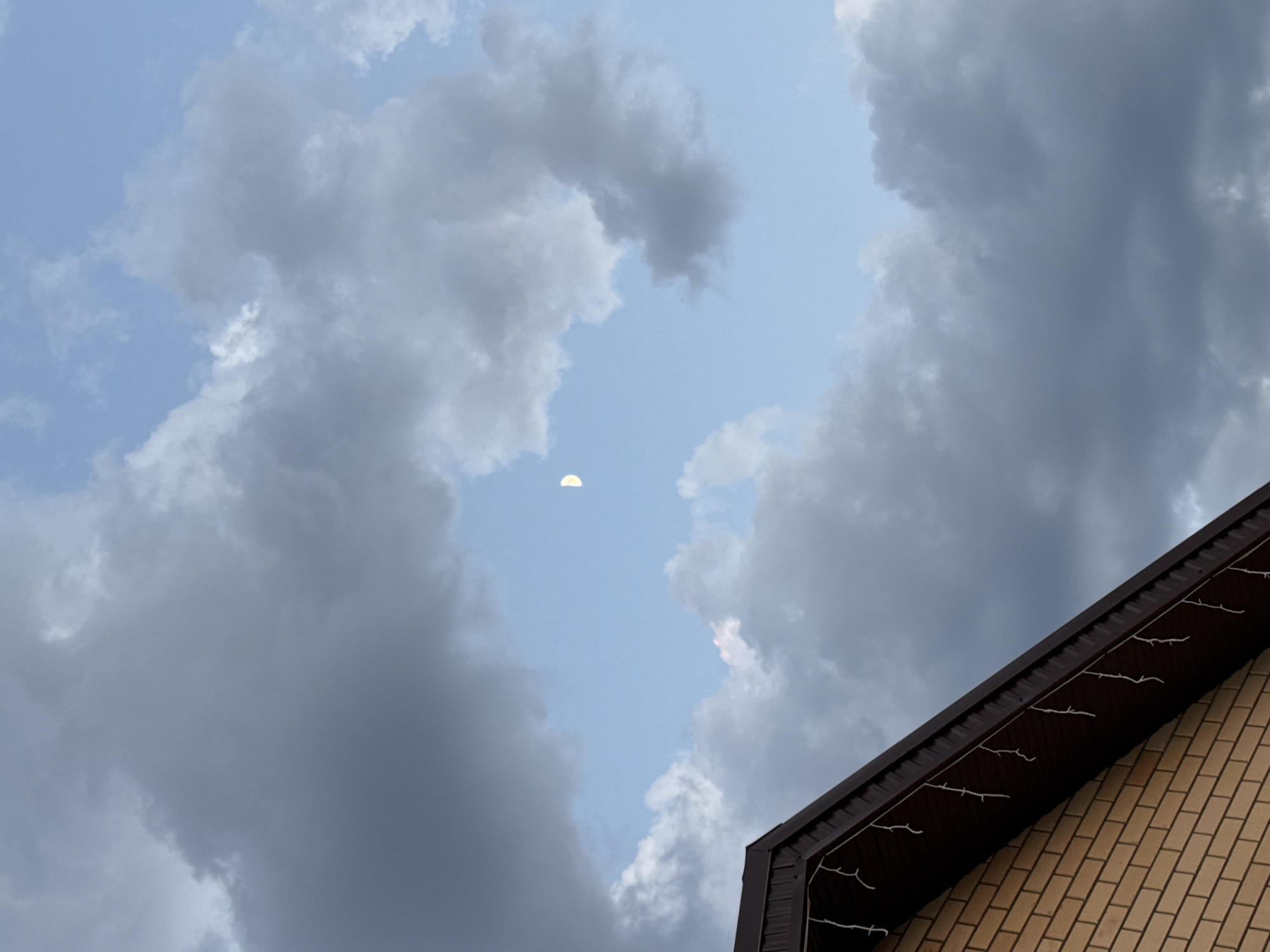 The image shows a view of the sky partially obscured by clouds and the corner of a brick building. In the sky, a crescent moon is visible through a gap in the clouds. The building's corner is made of light-colored bricks, and a dark brown roof overhang is attached, decorated with white string lights. The clouds range in color from light gray to dark gray, suggesting varying density. The sky is light blue. The location is inferred to be Ферма, Russia, based on external context.