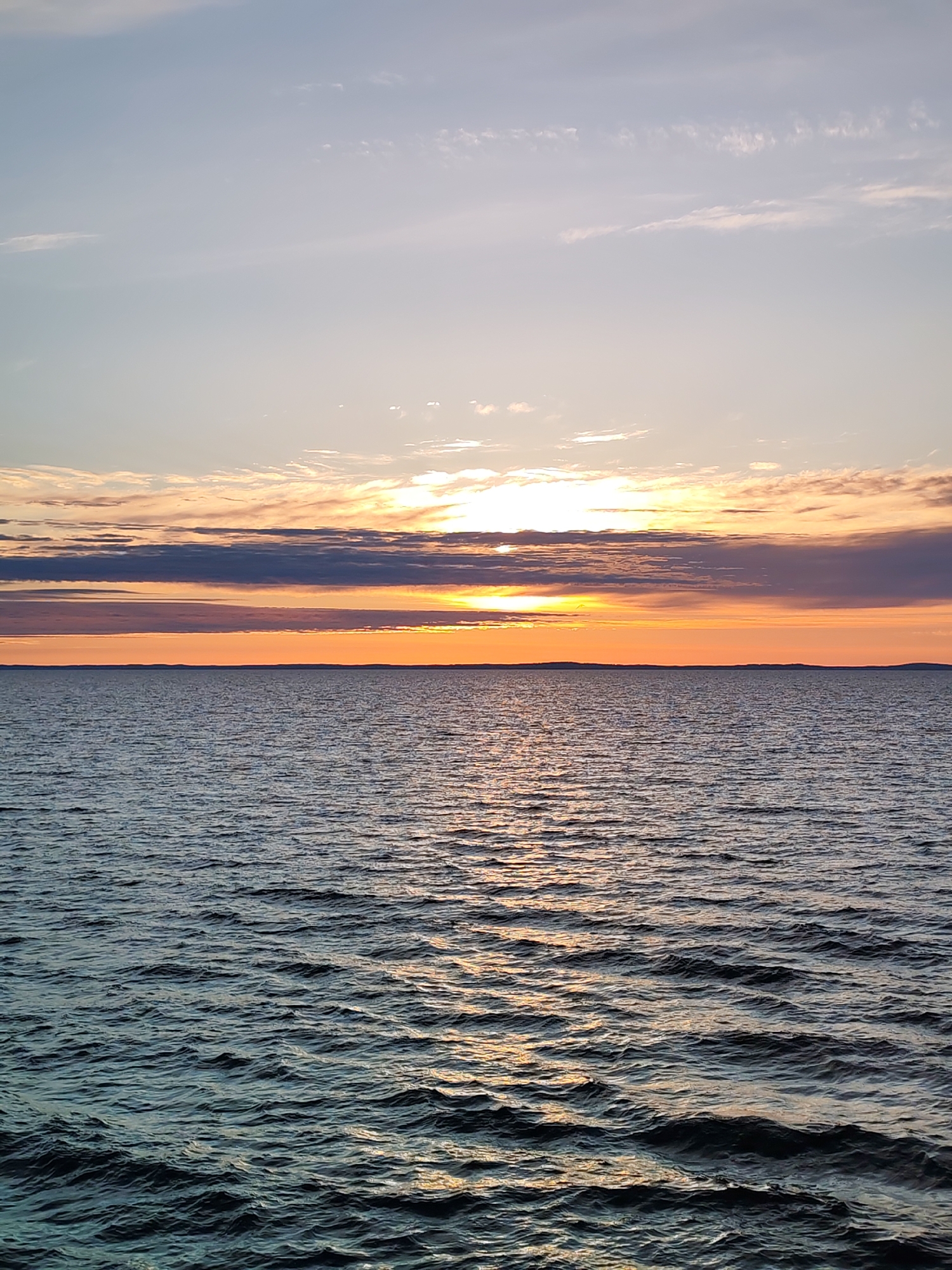 This media file captures a panoramic view of a tranquil sunset or sunrise over a vast body of water, likely a large lake or sea, extending towards a distant, flat horizon. The sky is divided into distinct zones of color and cloud formations. The uppermost section of the sky is a pale, clear blue with a few scattered, wispy clouds. Below this, a broad band of clouds stretches horizontally, displaying a range of colors from dark grey and purple-blue to vibrant orange and yellow, intensified by the sun's direct light.
The sun, a brilliant golden disc, is partially obscured by clouds just above the horizon, casting a strong, radiant glow. Directly beneath this bright area, the sky transforms into a vivid, intense orange and reddish hue, creating a striking contrast with the darker clouds above and the water below. A faint, dark silhouette of distant land or coastline is visible along the horizon line.
The surface of the water is characterized by gentle ripples and small waves, reflecting the colors of the sky, particularly the golden and orange light from the sun, which appears as shimmering streaks across the dark, blue-green water. There are no people, boats, or man-made objects visible in the frame, emphasizing the natural beauty and solitude of the scene. The image evokes a sense of peace and natural grandeur. The weather appears fair and calm. No text is visible in the image. This scene is set in an unknown location within Russia.