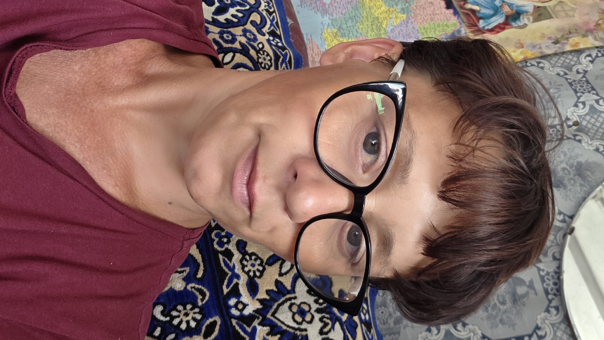 A close-up selfie of a woman wearing black cat-eye glasses and a burgundy t-shirt is shown. The woman has short, dark brown hair and fair skin. She is looking directly at the camera with a neutral expression. The background includes a tapestry or fabric with a blue and white floral pattern and a map with various colors. The scene appears to be indoors, possibly in a residential setting. The provided location of Vozsiiatske, Ukraine, is noted, though there are no specific visual cues within the image to confirm this location.