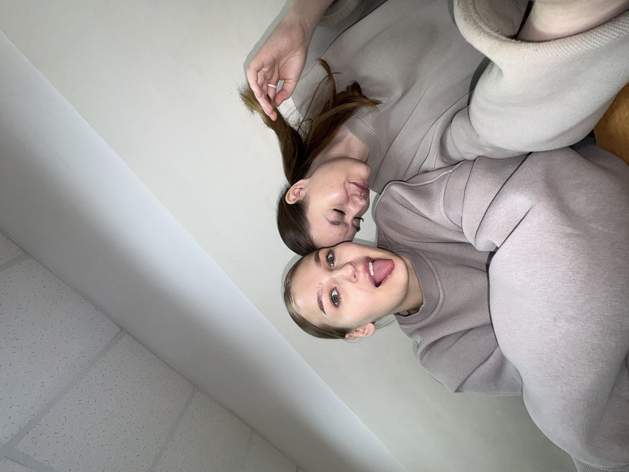 Two young women are taking a selfie from a low angle. The woman on the left is sticking her tongue out and looking at the camera. The woman on the right is leaning her head on her hand and playing with her hair. They are both wearing oversized sweatshirts and appear to be indoors. The background consists of a textured ceiling and a plain wall. The location is not specified in the image.