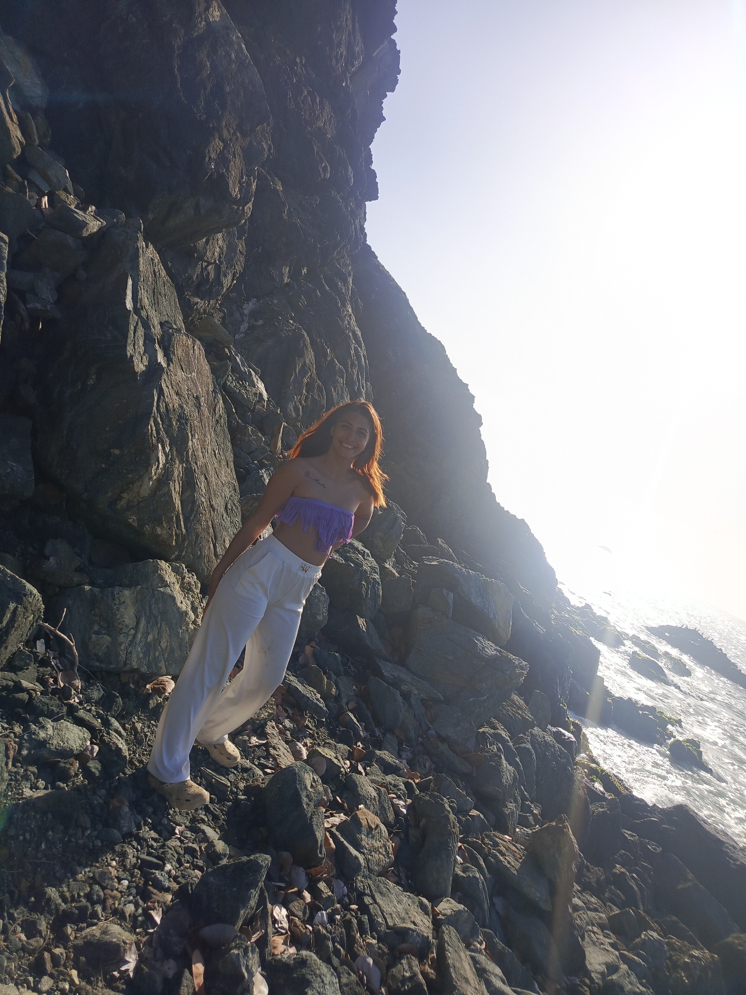 A woman with long, reddish-brown hair stands on a rocky incline. She is wearing a purple, fringed bandeau top and loose-fitting white pants. The woman is smiling and looking towards the camera. She is positioned on the left side of the image, leaning slightly against a large rock face. The background shows the ocean with sunlight reflecting off the water. The terrain is rugged and rocky, with scattered dry leaves on the ground. The scene is outdoors with bright daylight.