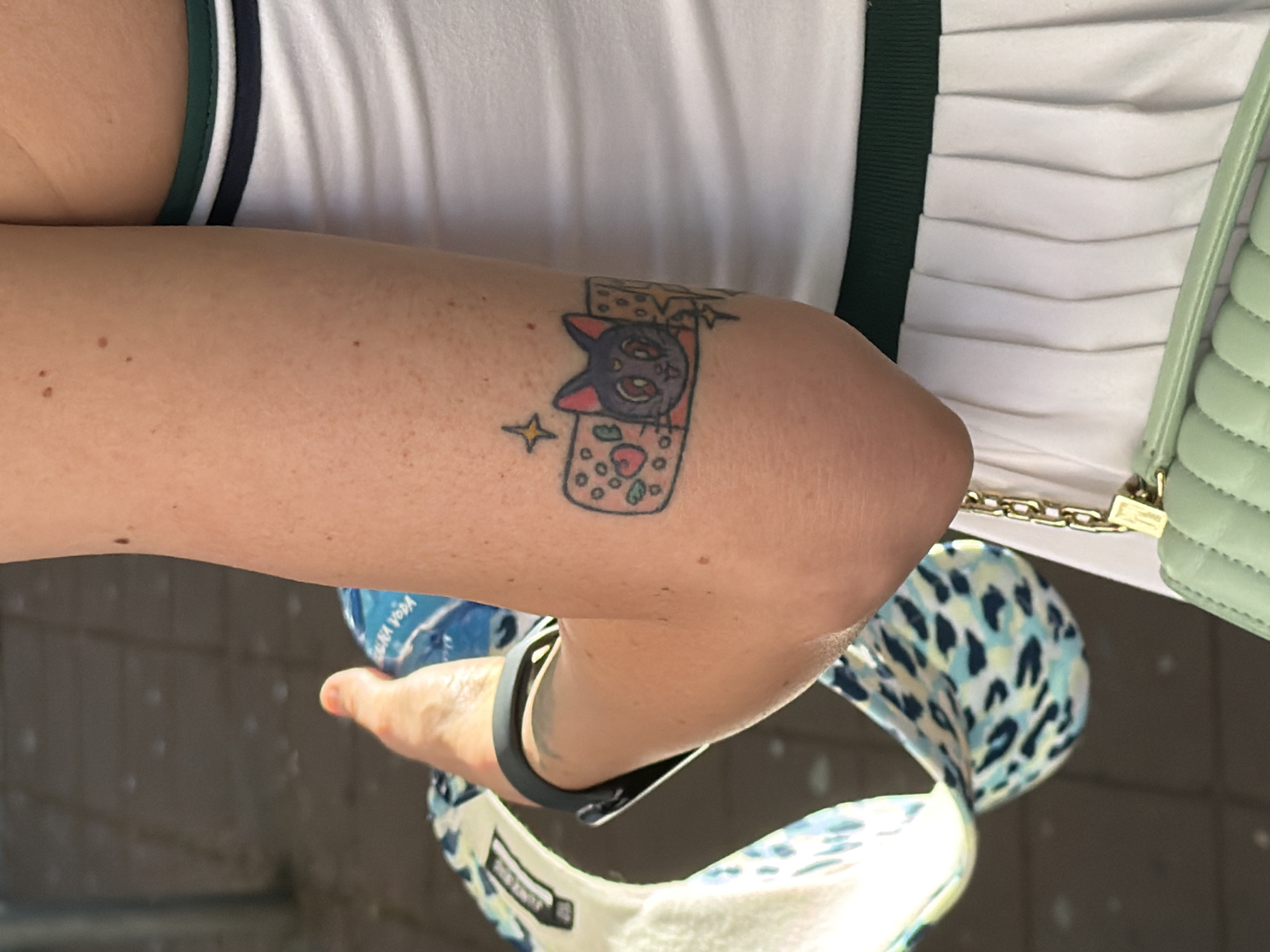 A woman stands outdoors with her left arm bent, displaying a tattoo on her upper arm. The tattoo features a cartoon cat face inside a band-aid adorned with small hearts and stars. The woman wears a white top with green and black trim. A light green quilted purse hangs at her side, and she carries a blue and white patterned bag. A black fitness tracker is on her wrist. A bottle of water is also visible in her hand. Based on the flooring tiles in the background, the location could be in a public outdoor space in Belgrade, Serbia.