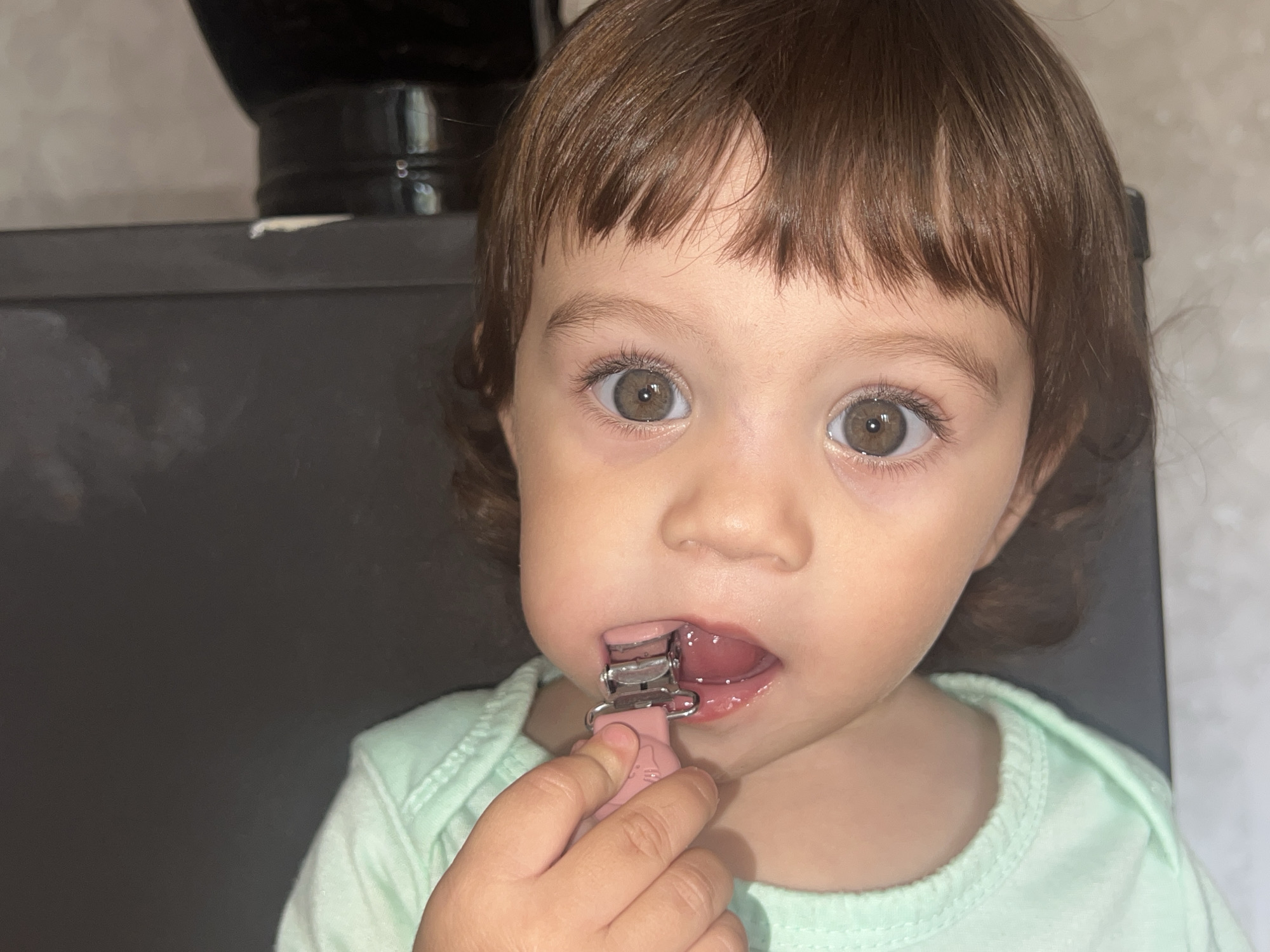 A close-up, eye-level shot shows a toddler with light brown hair and large, light brown eyes looking directly at the camera. The toddler is wearing a light green shirt. The toddler's mouth is open, and a pink pacifier clip with a silver metal clamp is attached to their lips. The background is out of focus, appearing to be indoors with dark and light gray surfaces. The lighting suggests it is daytime. The overall scene conveys innocence and a candid moment of childhood. There is no visible text in the image. The location context is Elena, Bulgaria.