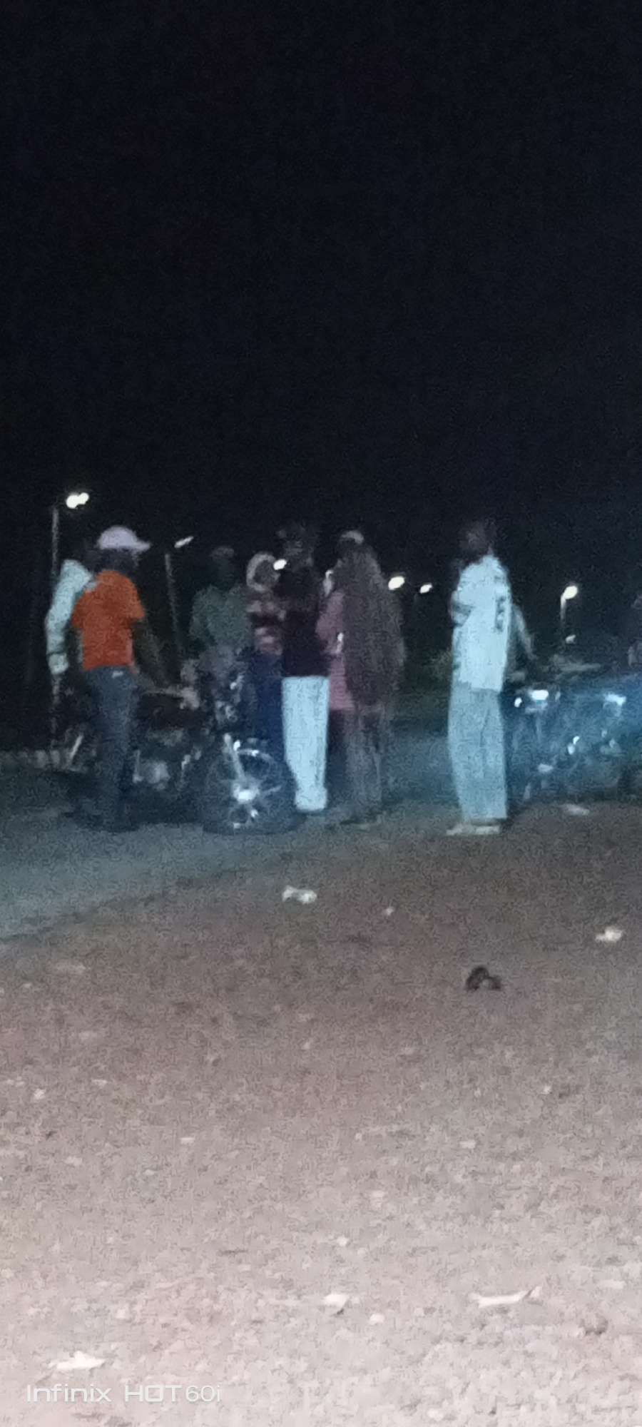 The scene is a poorly lit area in Adankolo, Nigeria, at night. A group of approximately seven people are clustered around at least two motorcycles. One person is wearing an orange shirt and light-colored hat. Another individual is wearing a pink top with long, dark hair. A person in white stands to the far right. Debris is scattered on the ground. Street lights are visible in the background.