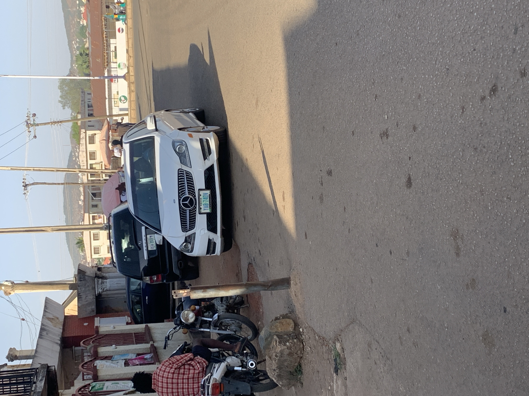 A street scene near the New Stadium in Nigeria. There are several vehicles parked along the side of the road: a white Mercedes-Benz, a black SUV, and two motorcycles. A person wearing a red and white plaid shirt is sitting on one of the motorcycles. The surrounding environment includes buildings and utility poles.