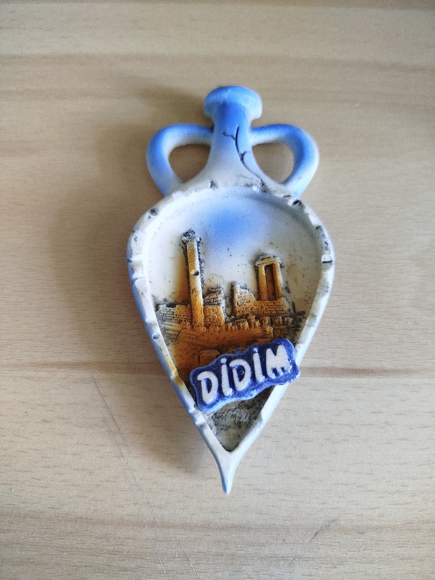 The image shows a tourist souvenir depicting ruins, possibly an ancient temple or historical site. The souvenir is shaped like a vessel, primarily white with blue accents. Inside the vessel-shaped souvenir are depictions of tan and orange colored structures, with a blue and white banner reading "DIDIM". The object rests on a light-colored wooden surface. The image provides no evidence to suggest a location in Varna, Bulgaria.