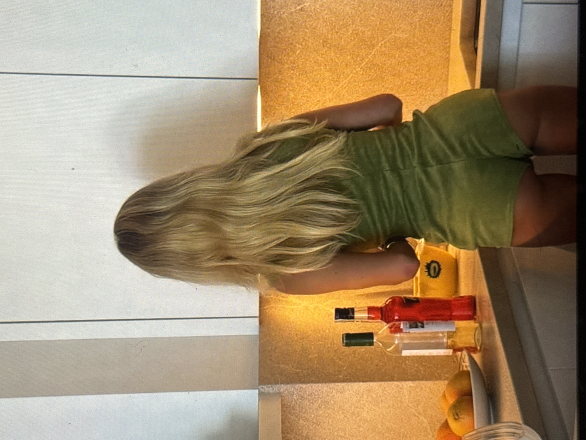 This media file depicts a woman with long, blonde hair, seen from the back. She is wearing a green outfit that appears to be a romper or a dress. The background is divided into two distinct sections. The left side features a white, vertically paneled surface. The right side is a textured, warm-toned surface that appears to be a wall or countertop, illuminated by warm light, suggesting it could be artificial lighting or the golden hour of daylight.
On the textured surface, several objects are visible. A yellow item with a black symbol resembling the number "0" inside a circle is present. Next to it, two bottles are placed horizontally. The top bottle is red, and the bottom bottle is clear with a light-colored label. Further to the right, a knife with a light-colored handle is positioned, and in the foreground, several round, orange fruits, likely oranges, are visible in what appears to be a glass container.
The lighting is warm and casts shadows, particularly on the white paneling on the left, indicating a light source coming from the right. The overall impression is an indoor setting, possibly a kitchen or a living space. The orientation of the image is such that the woman appears to be standing or leaning. There is no explicit information about the time of day, weather, or specific emotions being conveyed. The location context provided (Kuta Selatan, Indonesia) suggests a tropical environment.