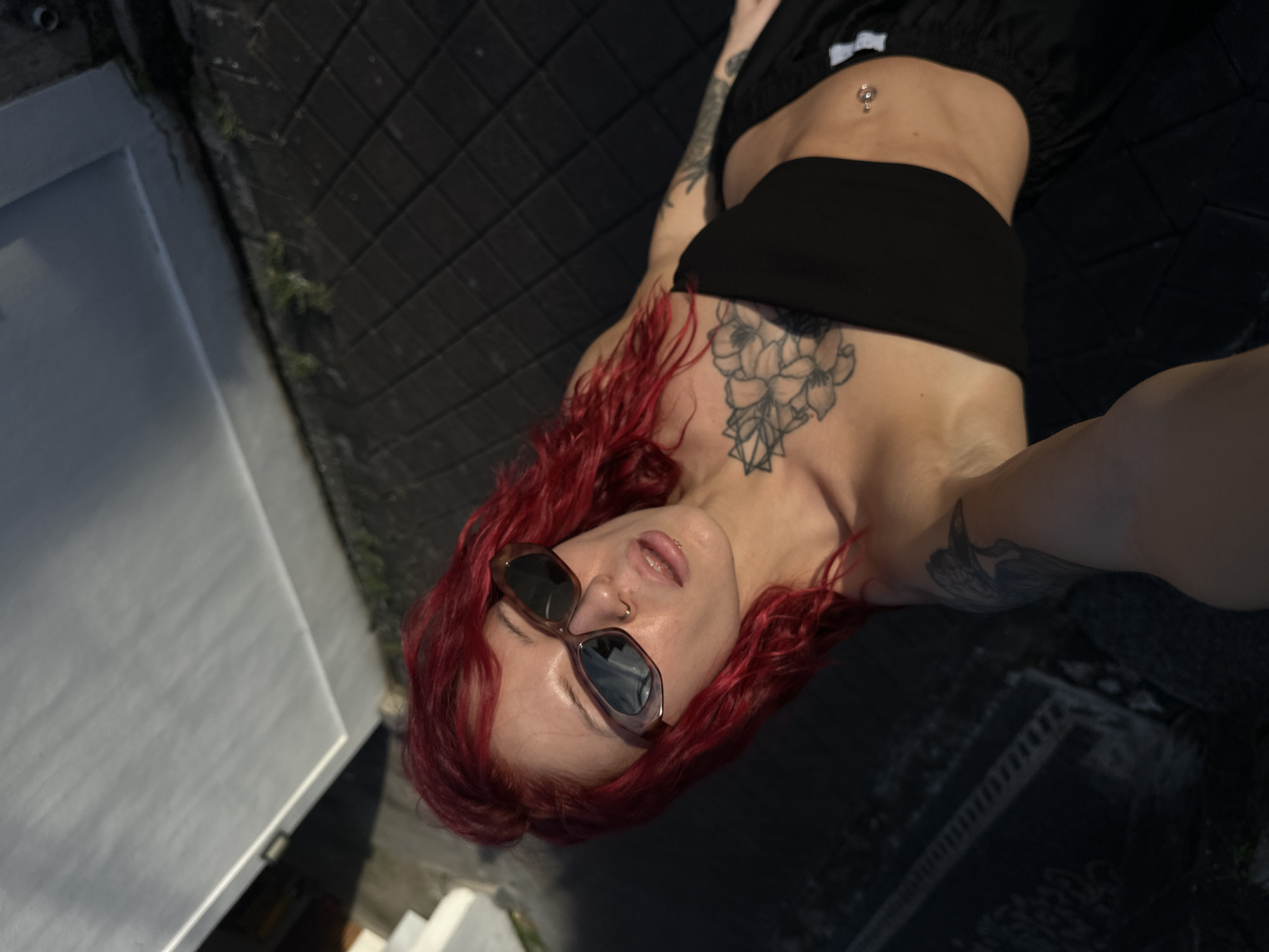 A woman with vibrant red hair is depicted in a high-angle image, likely a selfie, taken outdoors in North Kuta, Indonesia. She is reclining on a dark, paved surface, possibly a patio or balcony, with light-colored architectural elements visible to the left. The woman wears sunglasses, a black bandeau top, and dark pants, revealing a bare midriff with a navel piercing. She also has a small nose piercing. Several tattoos adorn her body, including a prominent floral and geometric design on her upper chest and shoulder, and another design on her right arm. Her lips are slightly parted, and she looks towards the camera with a relaxed expression. Bright daylight illuminates the scene, casting strong shadows and indicating clear, sunny weather. Small patches of green vegetation are visible along the edges of the paved area. There is no visible text in the image.