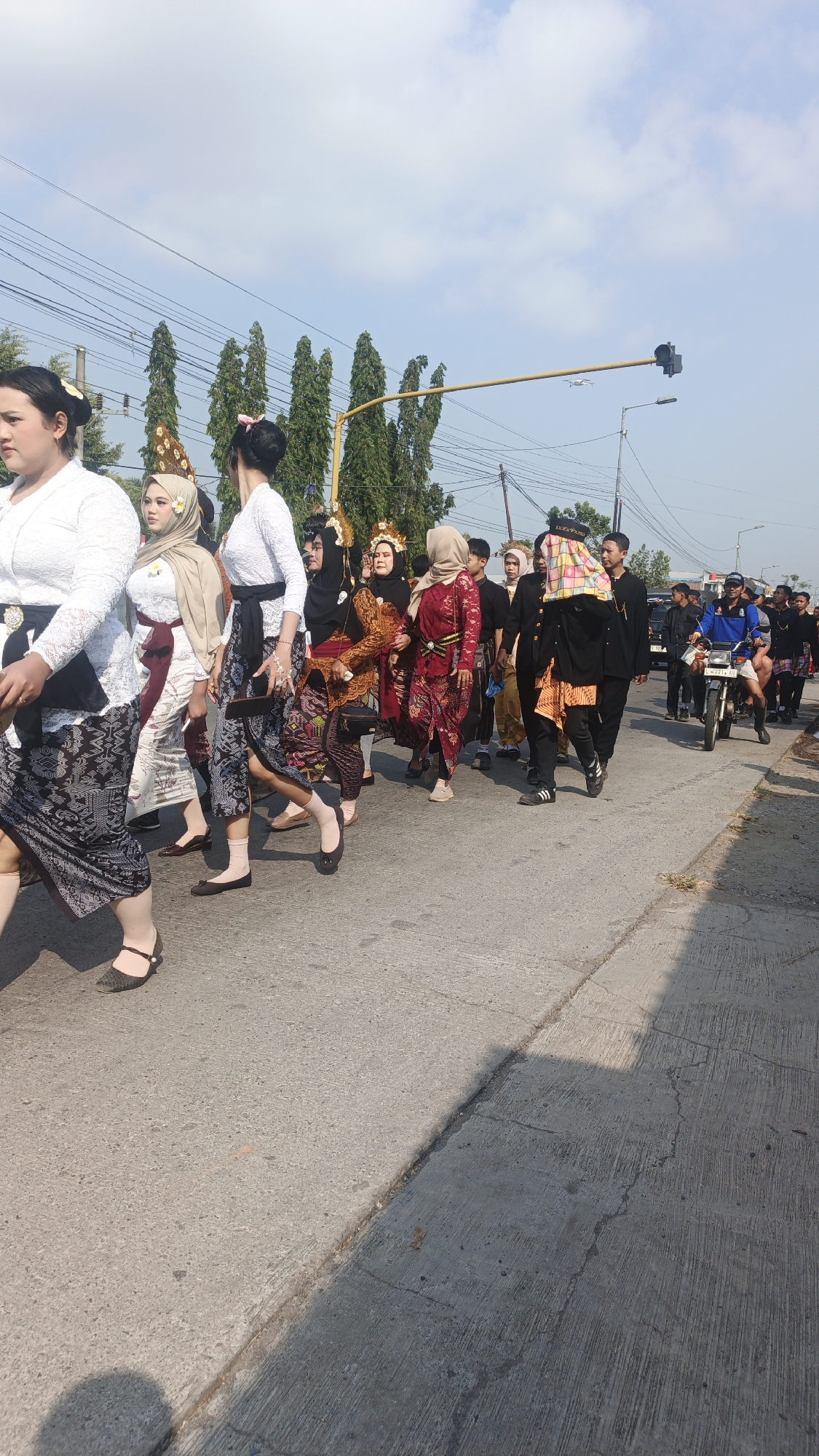 A group of people in traditional attire are walking along a paved road under a bright sky with scattered clouds. The procession appears to be in Sumengko, Indonesia, given the context. Several individuals are wearing colorful, ornate garments, including sarongs and headscarves. Some women have their hair styled in elaborate updos, adorned with flowers and decorative headpieces. A man in the foreground on the right is riding a motorcycle, leading the procession. In the background, there are trees, power lines, and a traffic signal. The road is bordered by a sidewalk on the right side, with sunlight casting shadows. The overall scene depicts a cultural event or parade taking place outdoors on a sunny day.