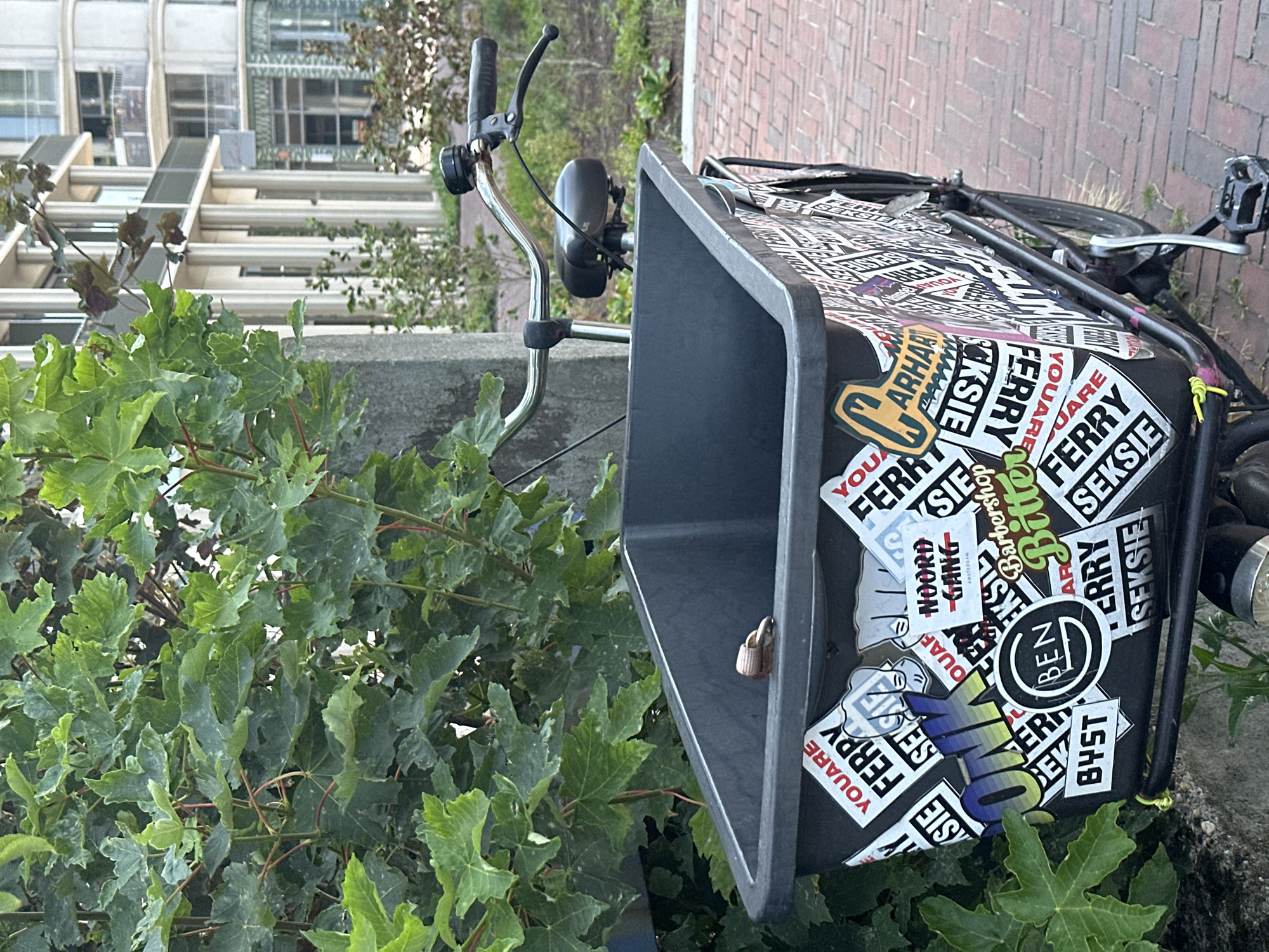 A black cargo bicycle is parked next to a concrete pillar and a leafy green bush. The bicycle's cargo box is covered in numerous stickers, many of which are white with black text. Some visible text includes "YOUARE FERRY SEXSIE", "ONK", "BEN", "NOORD GANG", "AMSTERDAM", "Barbershop", and "Bitter". The bicycle has a silver handlebar with a black grip and brake lever, and a black seat. In the background, modern buildings with white columns and balconies are visible. The ground is paved with red brick. The scene appears to be in a city, likely Amsterdam given the "AMSTERDAM" sticker.