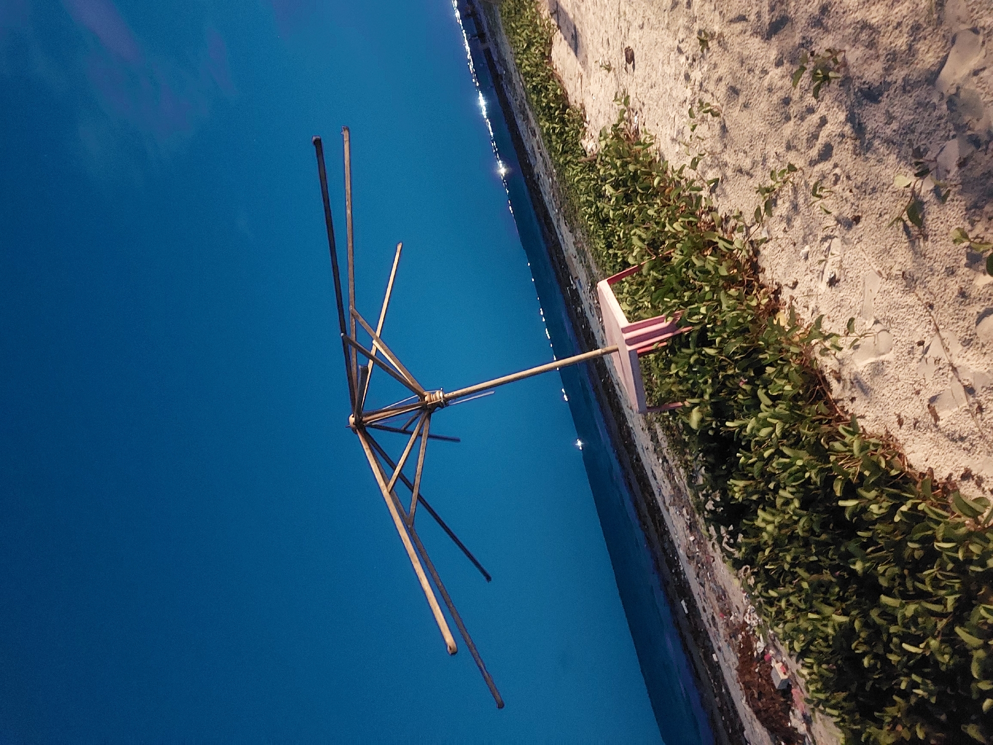 An unfolded clothesline, composed of a central pole and radial metal arms, stands adjacent to a seawall and a sandy beach covered with greenery. The background features a deep blue sky or water body, possibly the Indian Ocean. Reflective points along the shoreline suggest lights in the distance. This scene is located near the coast of Dar es-Salaam, Tanzania.