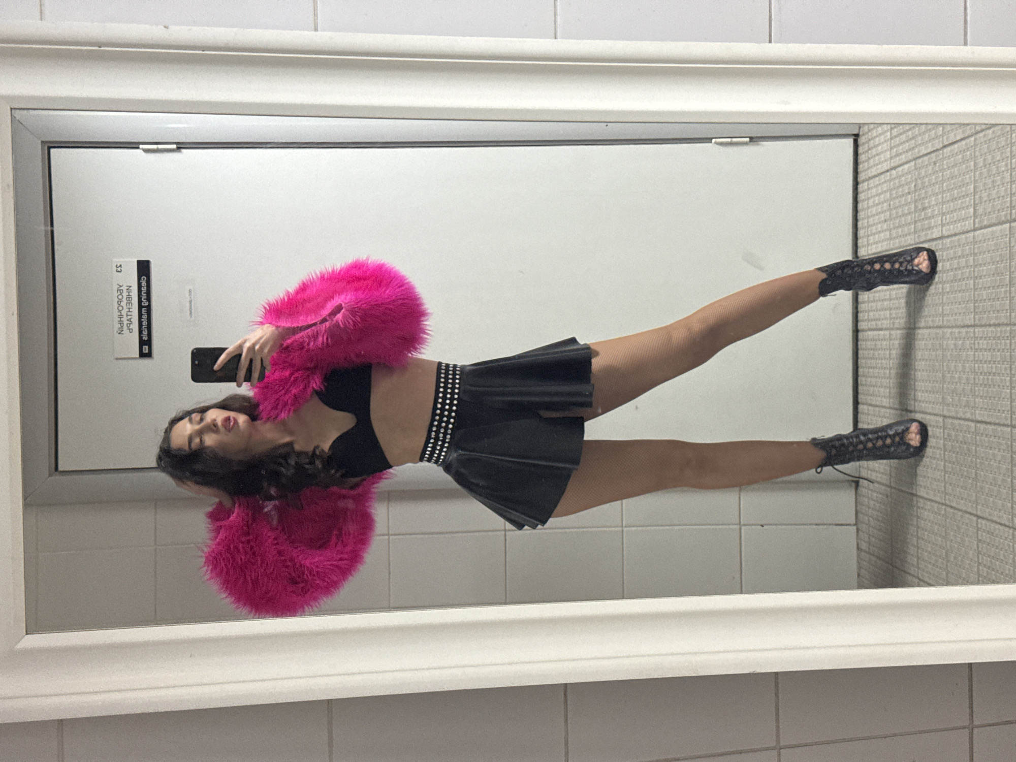 A woman is taking a mirror selfie in what appears to be a public restroom stall. She is wearing a black bra, a black mini-skirt with a studded belt, a bright pink fluffy jacket, and black open-toe heeled boots. Her reflection shows her holding a black smartphone in her right hand. The stall has tiled walls and floor. A sign is visible on the door behind her, with text that reads "CLEANING ENSUED" in English. The door also has a small metallic plaque with text that appears to be in a Slavic language. The overall impression is that of a private moment captured in a utilitarian space, possibly in Kazan, Russia, given the context of the sign's text, though this cannot be definitively confirmed from the image alone.