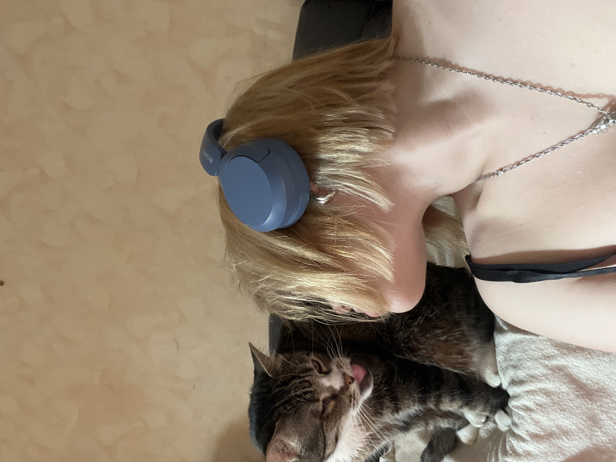 This media file presents an indoor scene depicting a person and a cat in a relaxed setting. The image is taken from an overhead perspective, with the subjects lying horizontally.
The main subject is a person, likely female, with short, light blonde hair. They are wearing blue-grey over-ear headphones, with the "SONY" brand name visible on the left earcup. A silver chain necklace is draped around their neck and chest. A black bra strap is visible on the right side of the frame, suggesting the person is wearing a bra or a similar top. Their skin appears smooth and light-toned.
Adjacent to the person, a tabby cat with brown, grey, and white fur is seen lying down. The cat's eyes are partially closed, and its pink tongue is slightly extended, suggesting it might be in the middle of grooming or simply very relaxed. Its paws are visible on a light-colored, textured blanket or sheet.
The background consists of a plain, light brown or tan wall with a subtle textured finish. The overall lighting is soft, indicating an indoor environment without harsh sunlight. The scene conveys a sense of calm and comfort. While no specific time of day is discernible from the image, the relaxed posture of both subjects suggests a leisure moment.
The visible text is "SONY" on the headphones. The location context provided is Odesa, Ukraine, though no specific visual cues within the image confirm this.