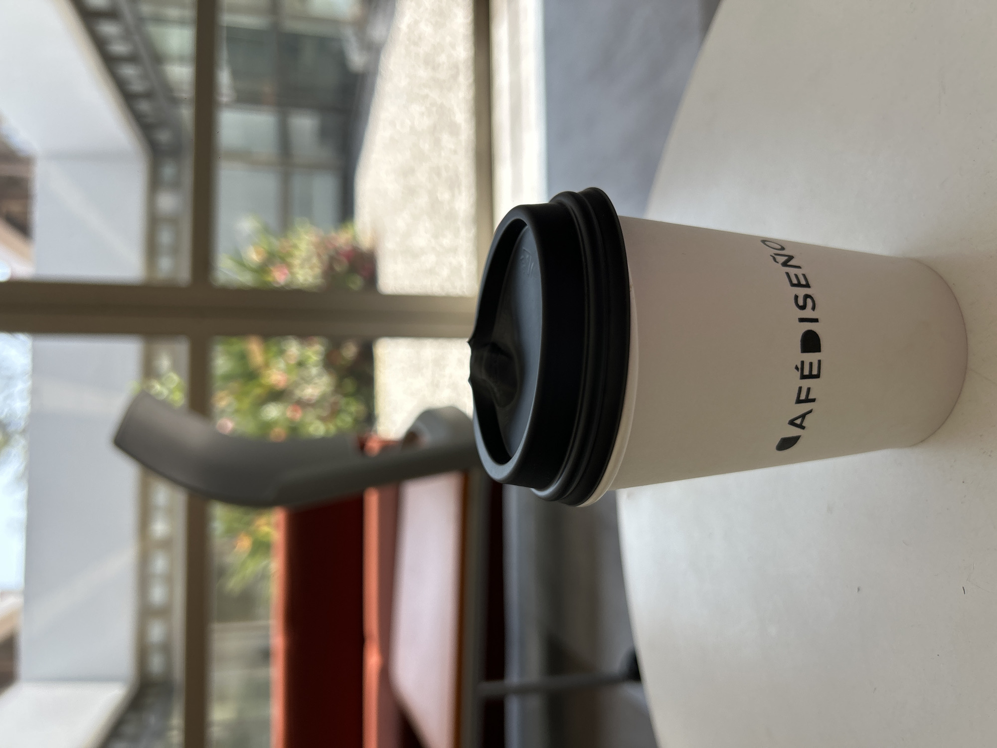 Here is a detailed description of the image:
The image shows a white paper coffee cup with a black lid on a white surface. The cup has the words "CAFÉ DISEÑO" printed vertically on it. The background is a blurred view of what appears to be the outdoors, seen through a window. The location appears to be indoors. A grey chair is placed behind the cup. The overall scene is a still life, capturing a moment possibly in a café or an indoor setting with a view to the outside.