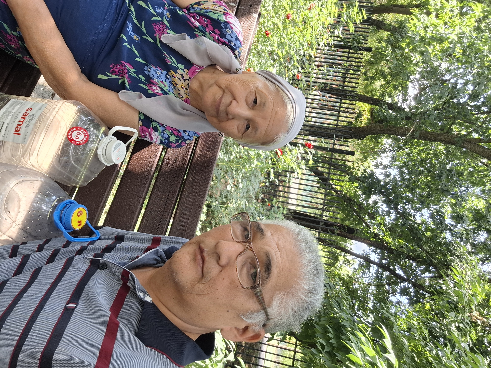 An elderly Asian woman and an elderly Asian man are seated on a wooden park bench. The woman is on the left, wearing a floral-print dress and a light-colored headscarf. She is looking towards the camera with a slight smile. The man is on the right, wearing a striped polo shirt and glasses. He is looking slightly off-camera. In front of them on the bench are two large plastic water bottles, one with a red and white label and the other with a blue and yellow cap. The background features a black metal fence, lush green trees, and some red flowers. The scene appears to be a park in Almaty, Kazakhstan, possibly during daylight hours, suggested by the bright sunlight filtering through the trees. The man is likely taking the selfie, as indicated by his position and gaze.